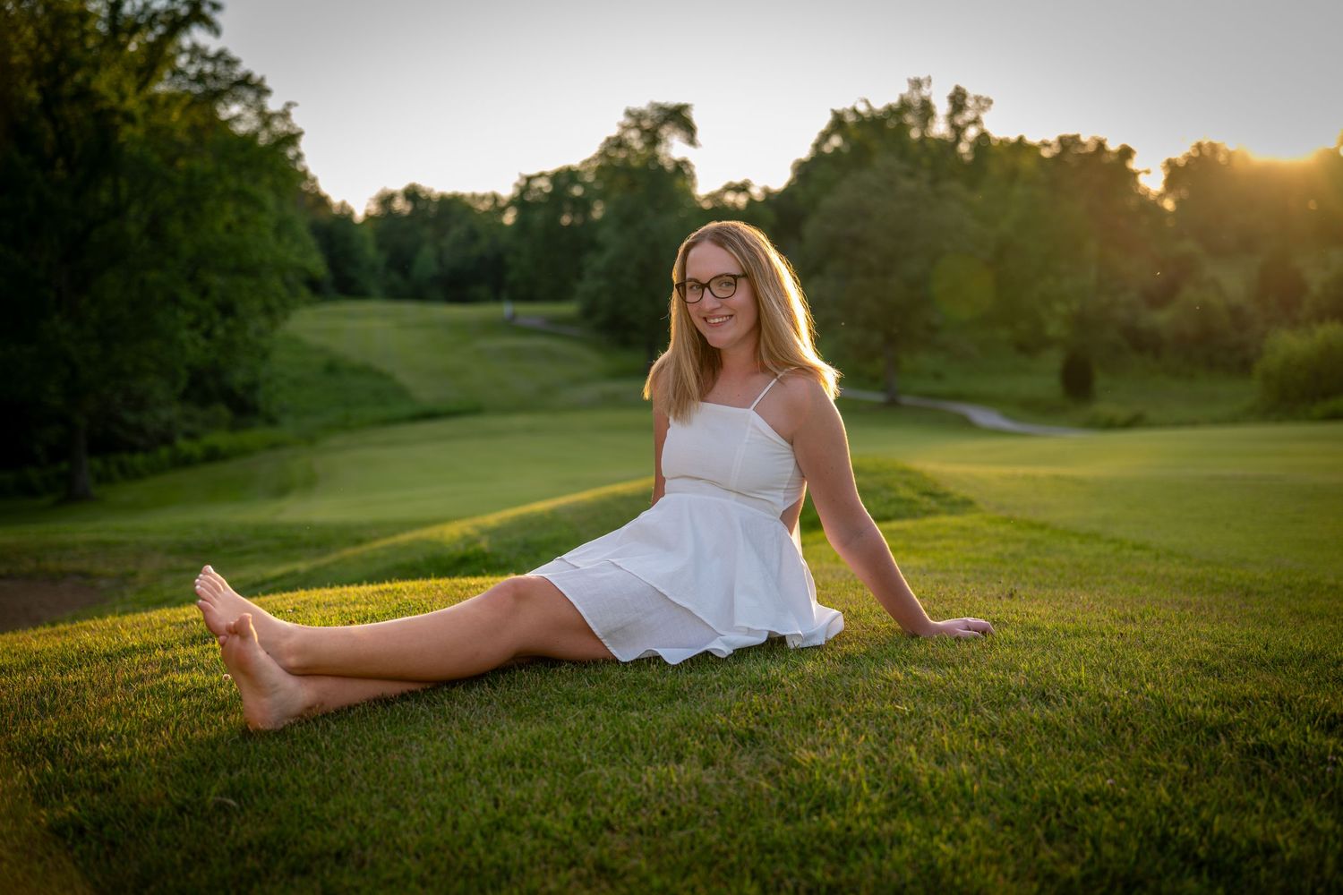 Smiling teenage girl in white dress sitting on lush green golf course grass at golden hour sunset.