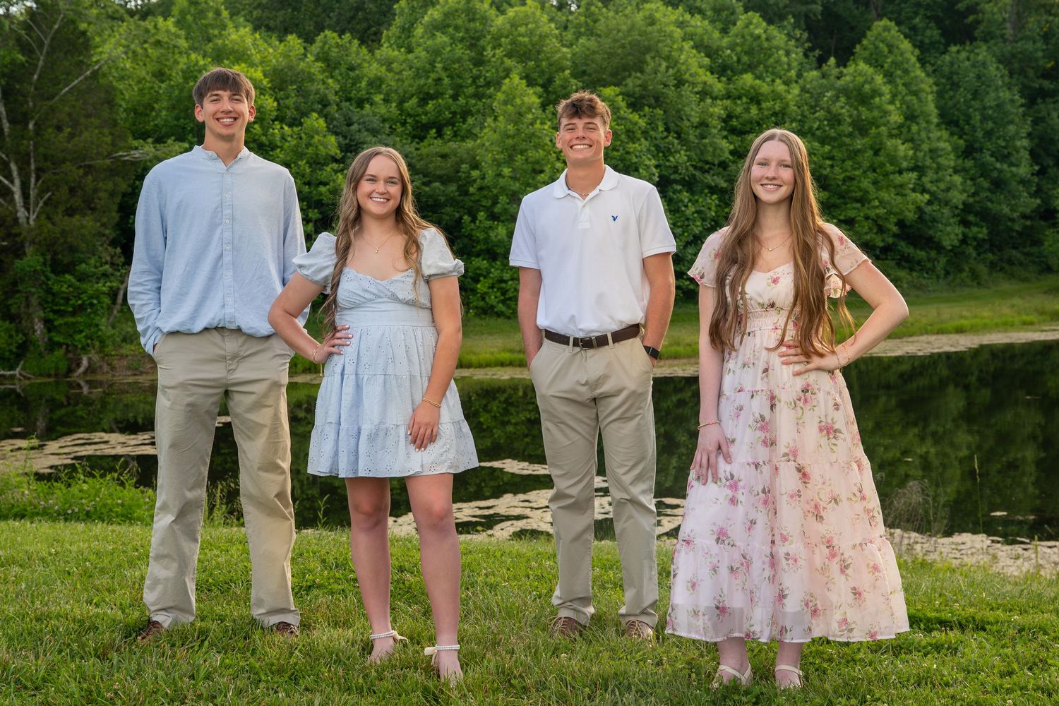 Four teenage students pose outdoors near a pond in semi-formal spring attire.