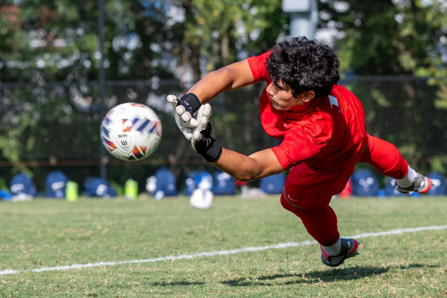 A soccer goalkeeper in red dives to make a save during a match on a green field.