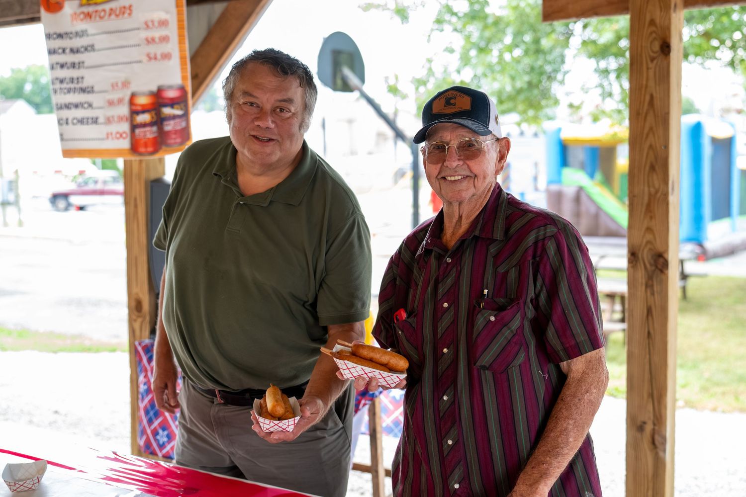 Two men smiling at a food stand, one holding a hot dog at an outdoor fair.