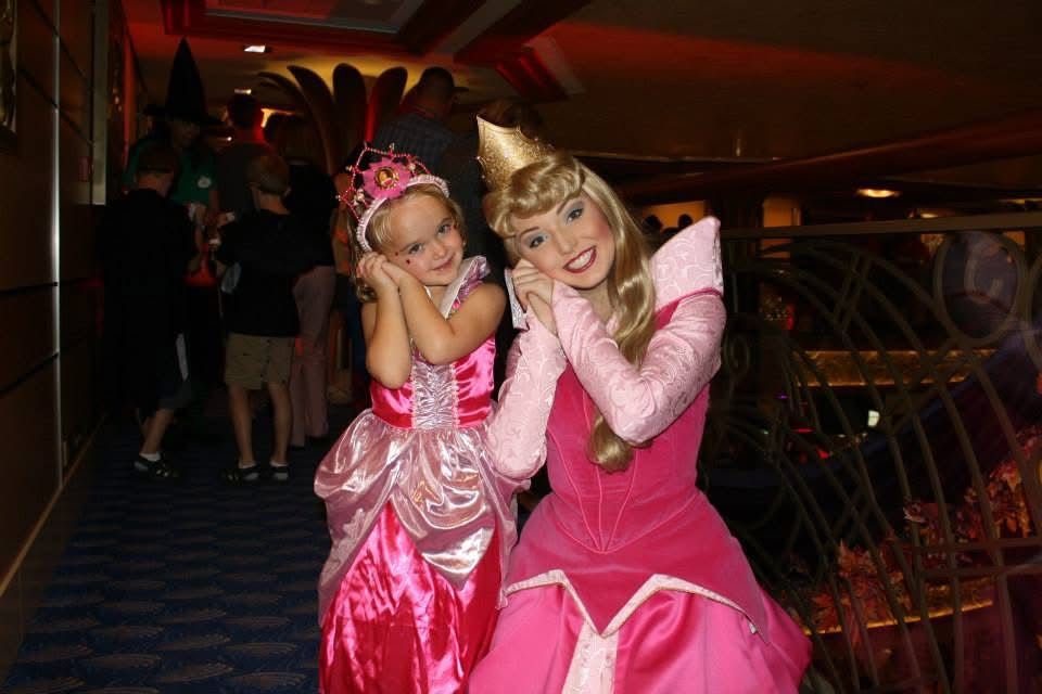 A young girl in a pink princess costume poses with a Sleeping Beauty character in a pink gown at a Disney event.