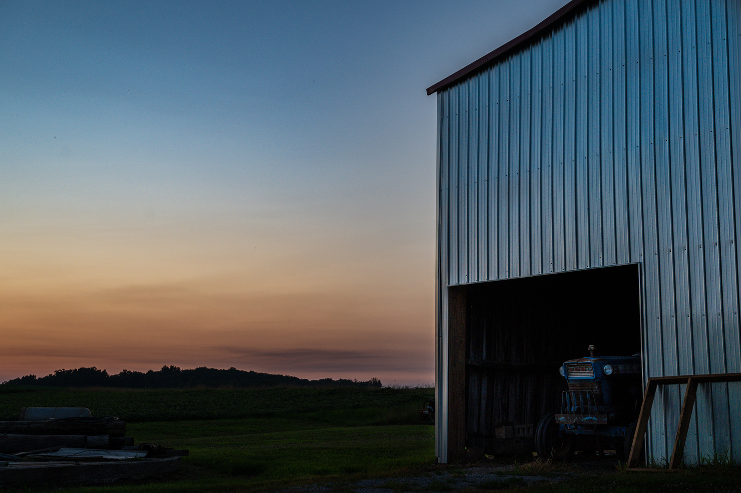 Metal barn at sunset with open door revealing equipment, overlooking misty green fields under a colorful dusk sky.