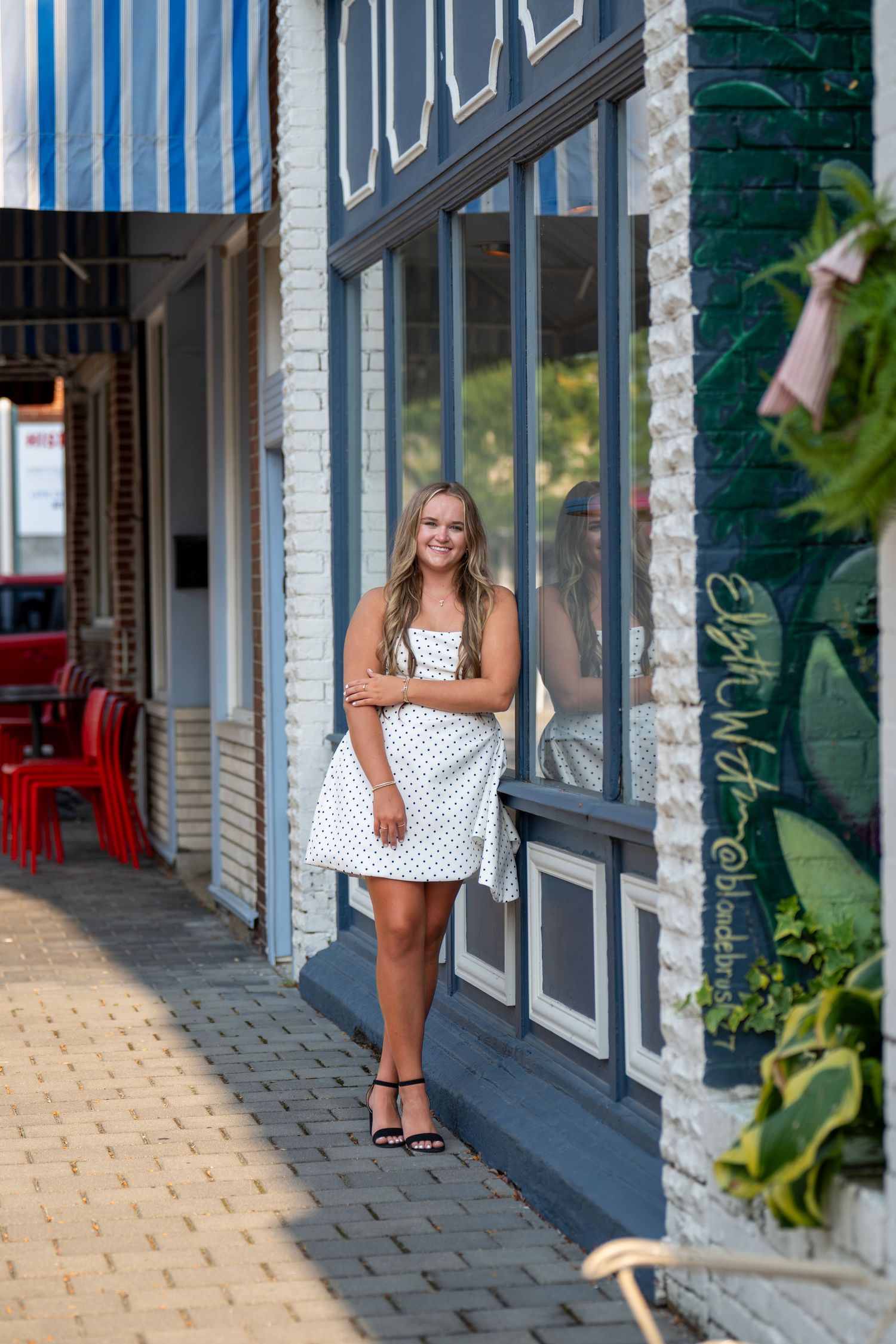 Smiling blonde teen in white polka dot dress poses outside boutique storefront on brick sidewalk.
