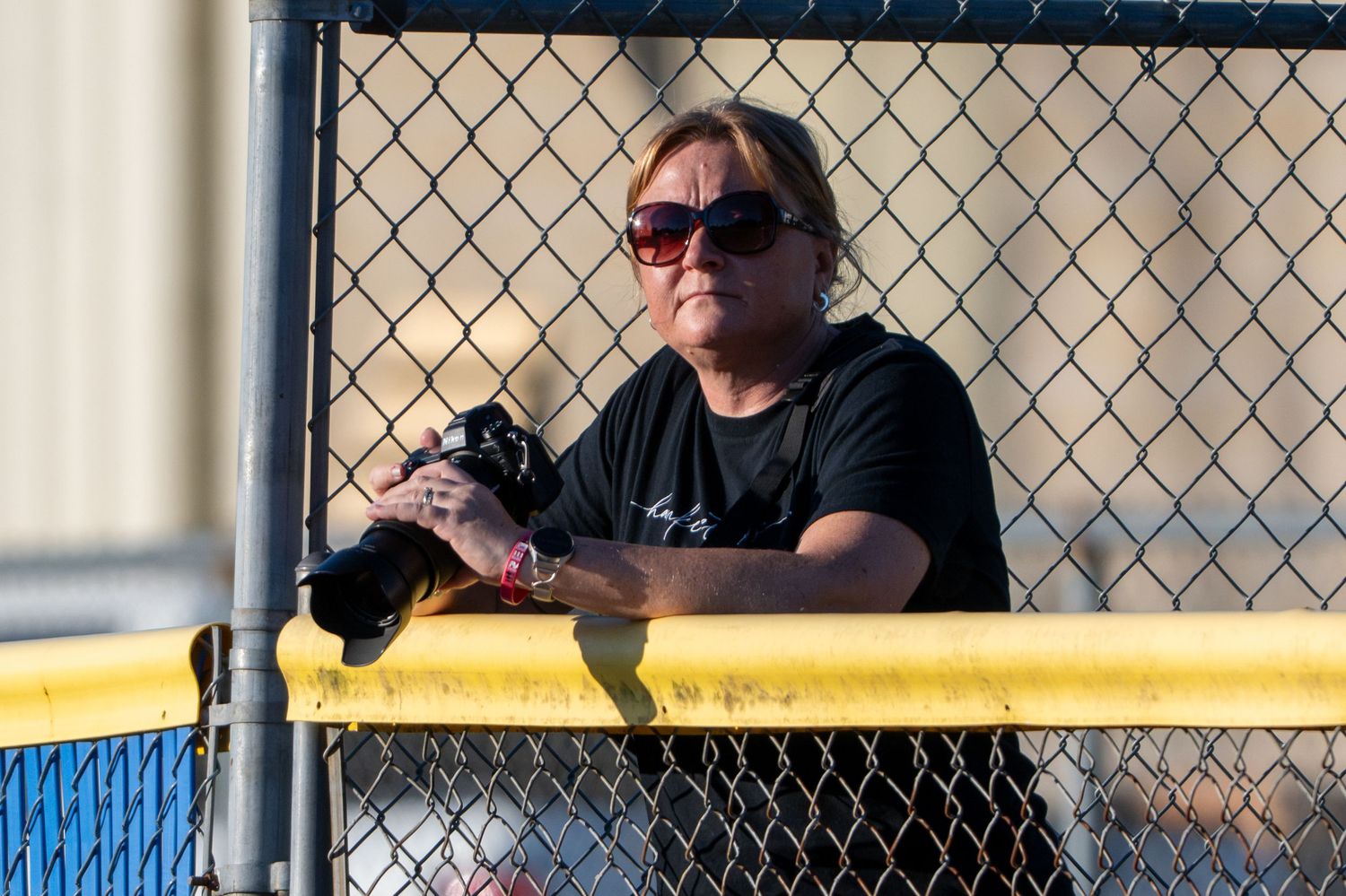Woman in black shirt and sunglasses holds camera while leaning on yellow fence at outdoor sports event.