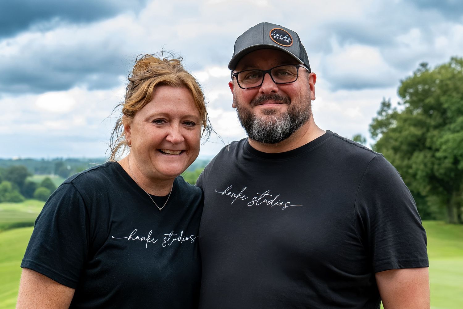 Smiling couple wearing matching black shirts outdoors on cloudy day.