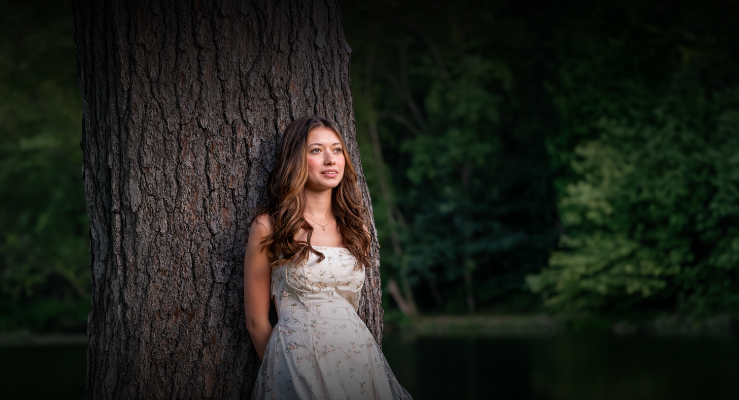 Young woman in elegant white dress leaning against large tree trunk with lush green forest background.