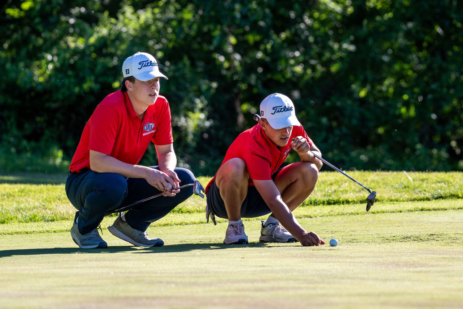Two Heritage Hills Patriots golfers in red shirts crouch to read the green before putting.