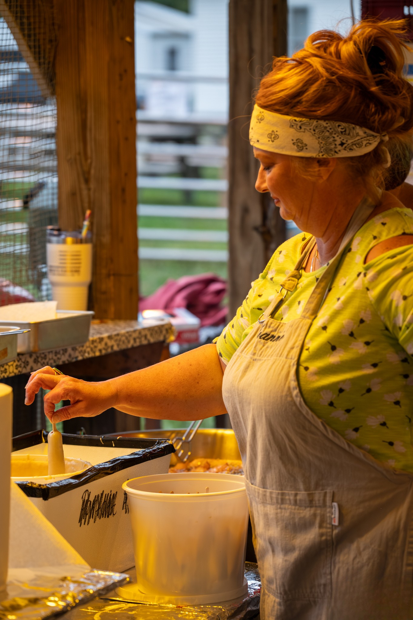 Woman in yellow floral shirt and apron cooking food at an outdoor kitchen station wearing a white headband.