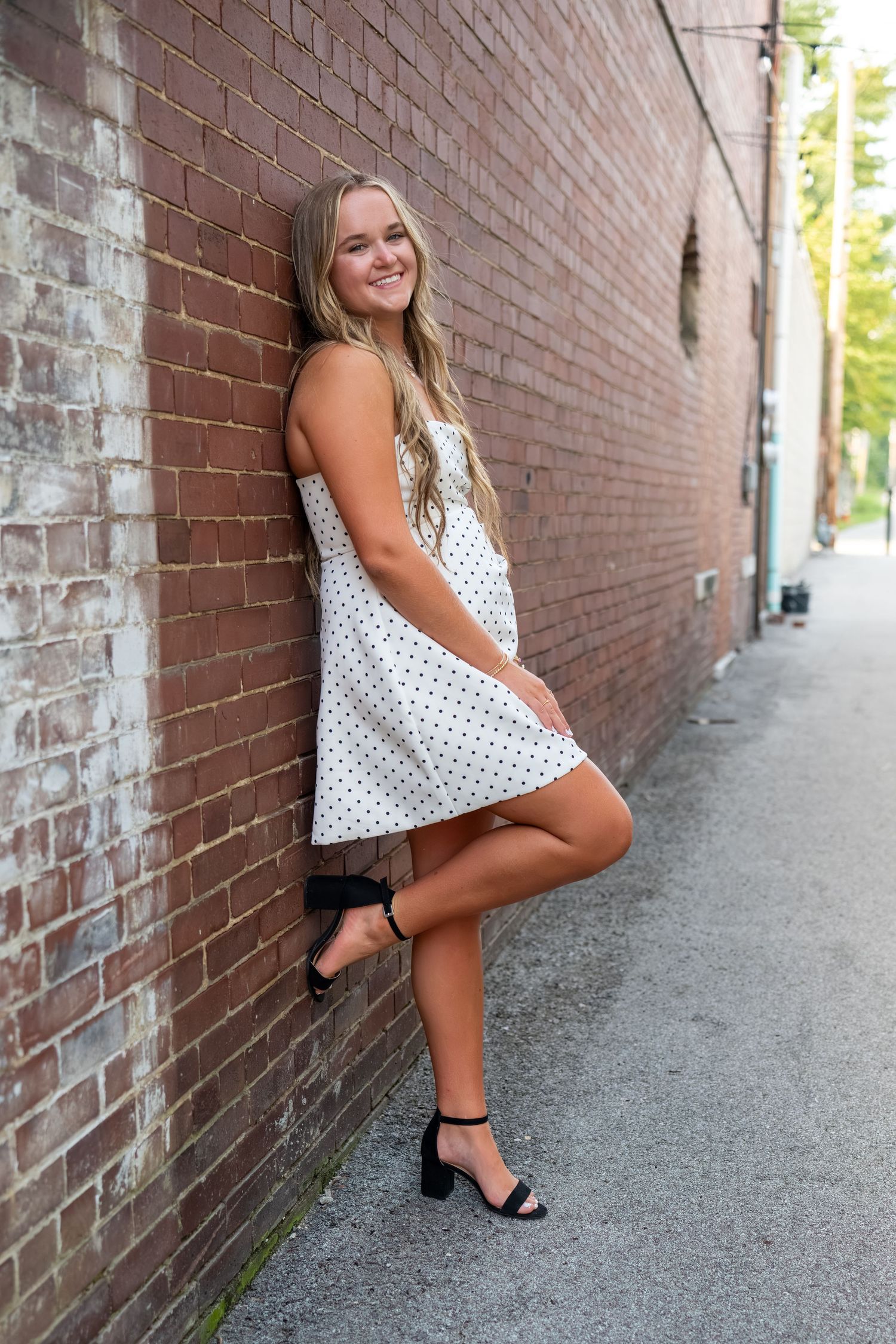 Young woman in white polka dot dress leaning against brick wall in urban alley wearing black heeled sandals.