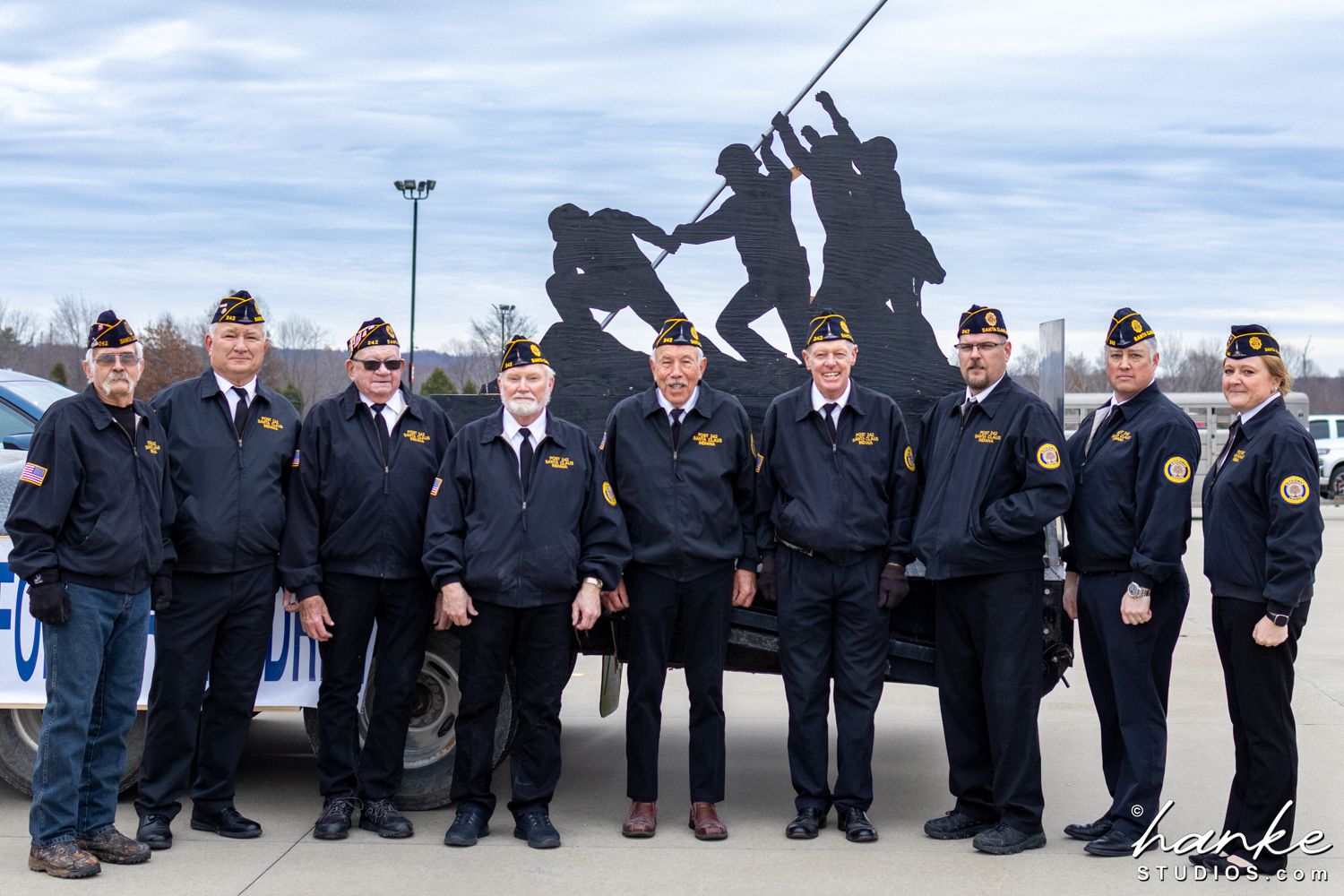 Group of veterans from the Santa Claus American Legion Post 254 posing in matching jackets before an Iwo Jima memorial sculpture outdoors.