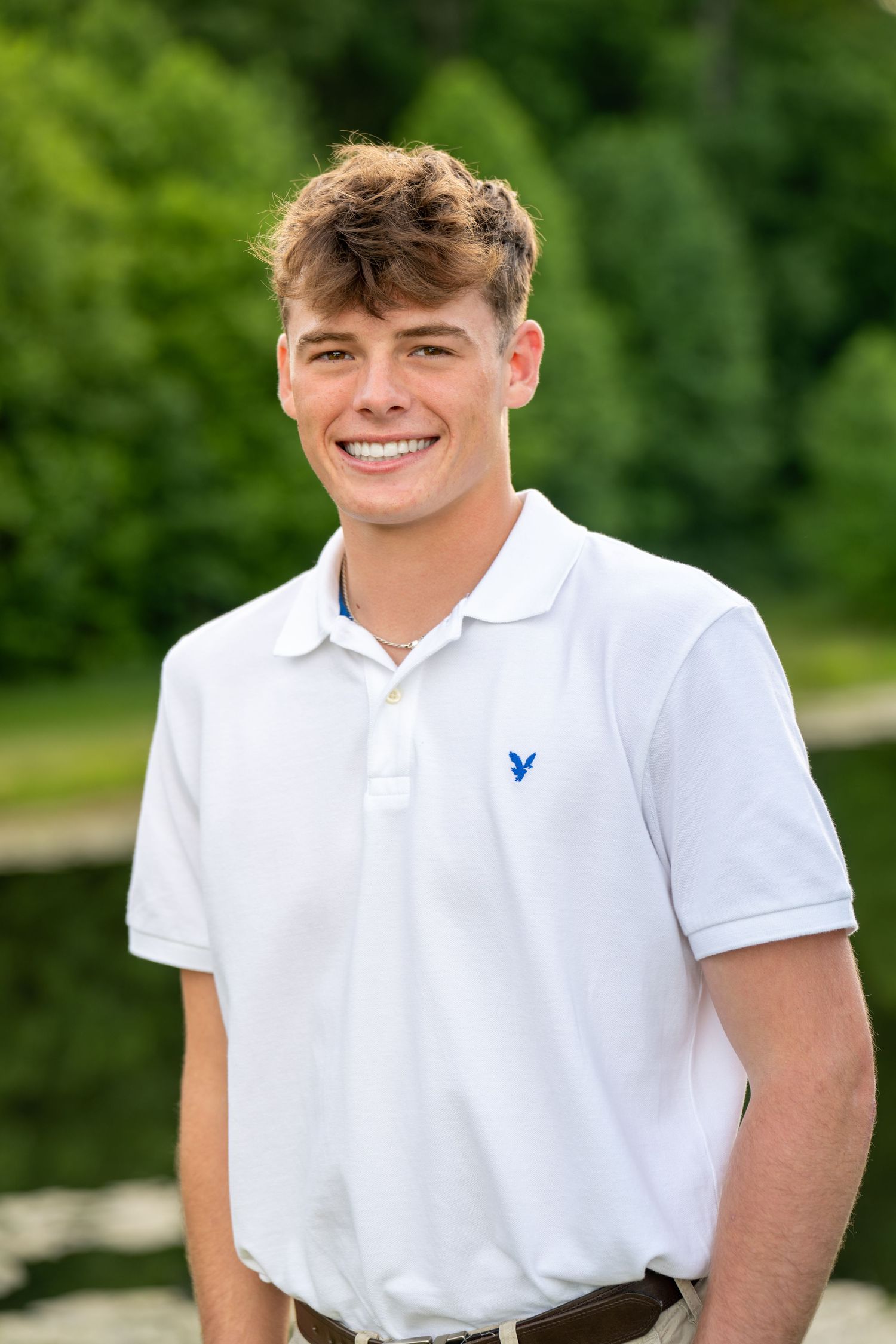 Smiling young man in white American Eagle polo shirt posing outdoors near pond with green trees.