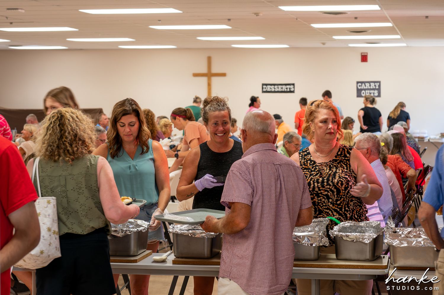 Community gathering in church hall with cross on wall, people serving food at buffet tables. Mariah Hill Picnic.