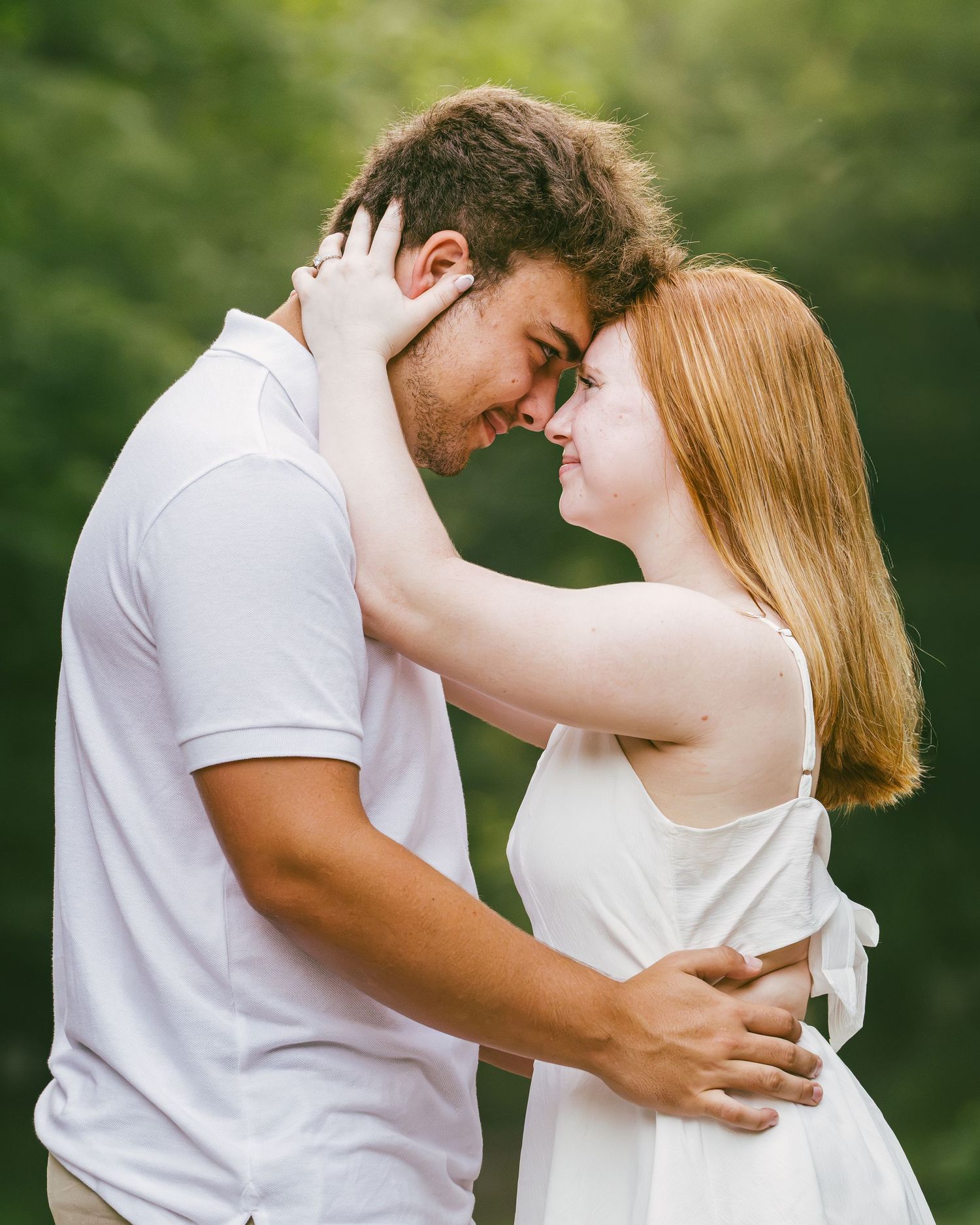 Romantic couple in white outfits sharing tender moment outdoors with green foliage background.