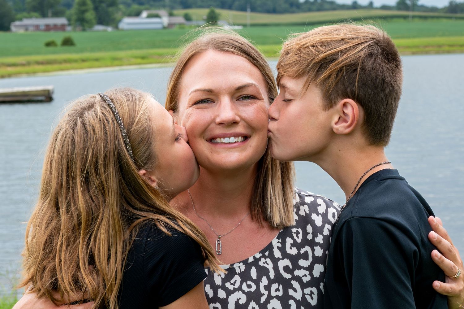 Happy blonde mother being kissed on cheeks by two children near a lake outdoors.
