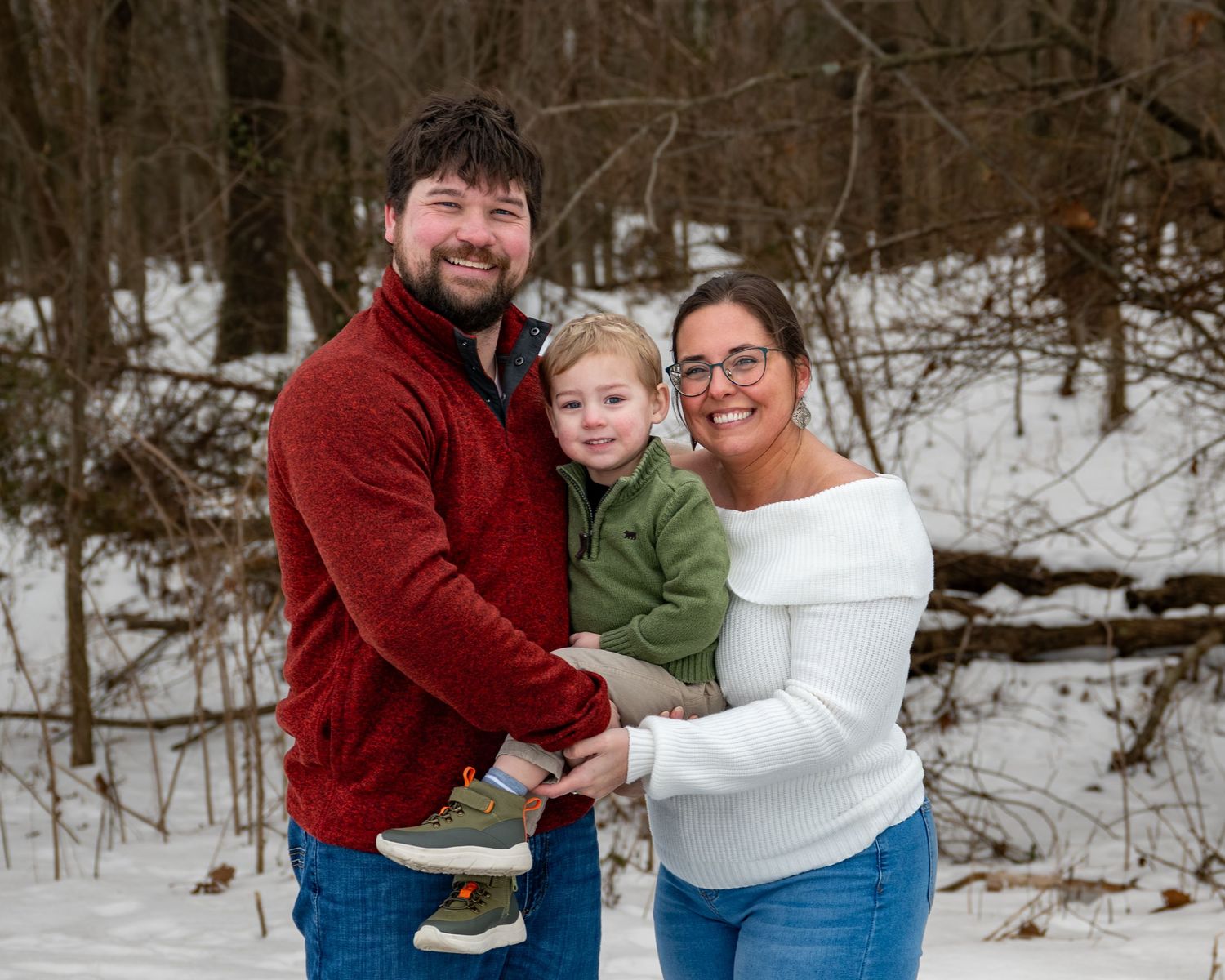 Smiling family of three posing together in a snowy winter forest setting.
