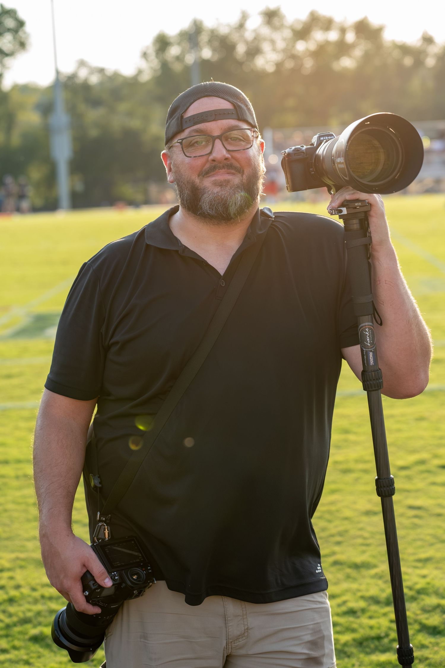 Bearded male photographer smiles holding camera and monopod on a sunny field.