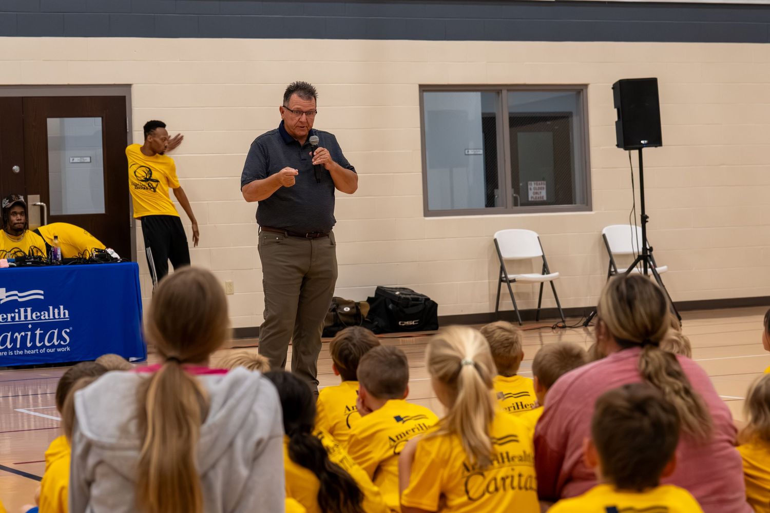 Senator Daryl Schmitt speaking into microphone to group of children in yellow shirts during AmeriHealth Caritas Healthy Hoops event at YMCA