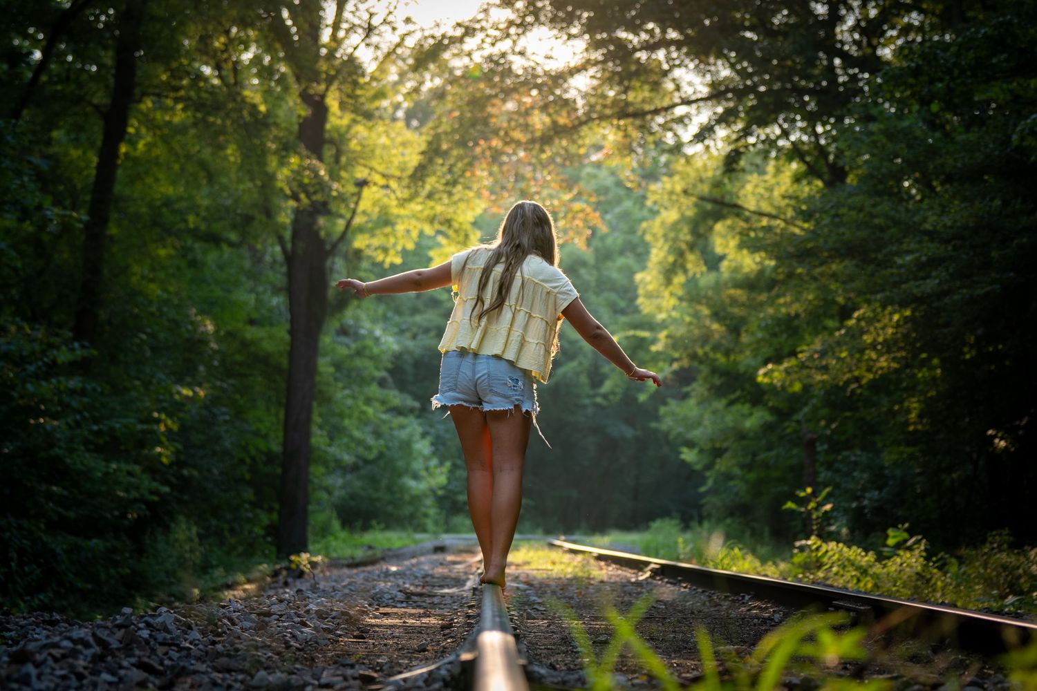 Young woman balancing on railroad tracks in a sunlit forest, arms outstretched, wearing denim shorts.