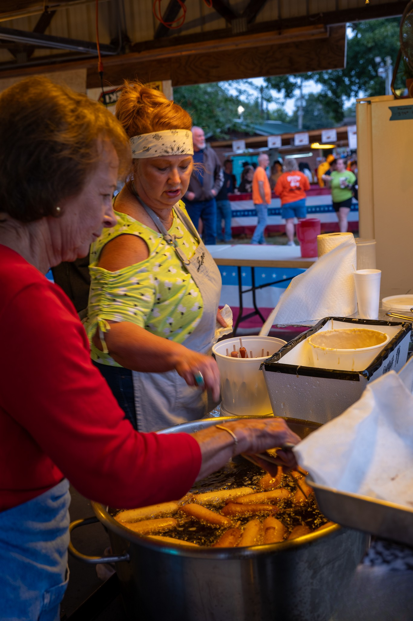 Two women serving fried food at an outdoor festival food booth at night.