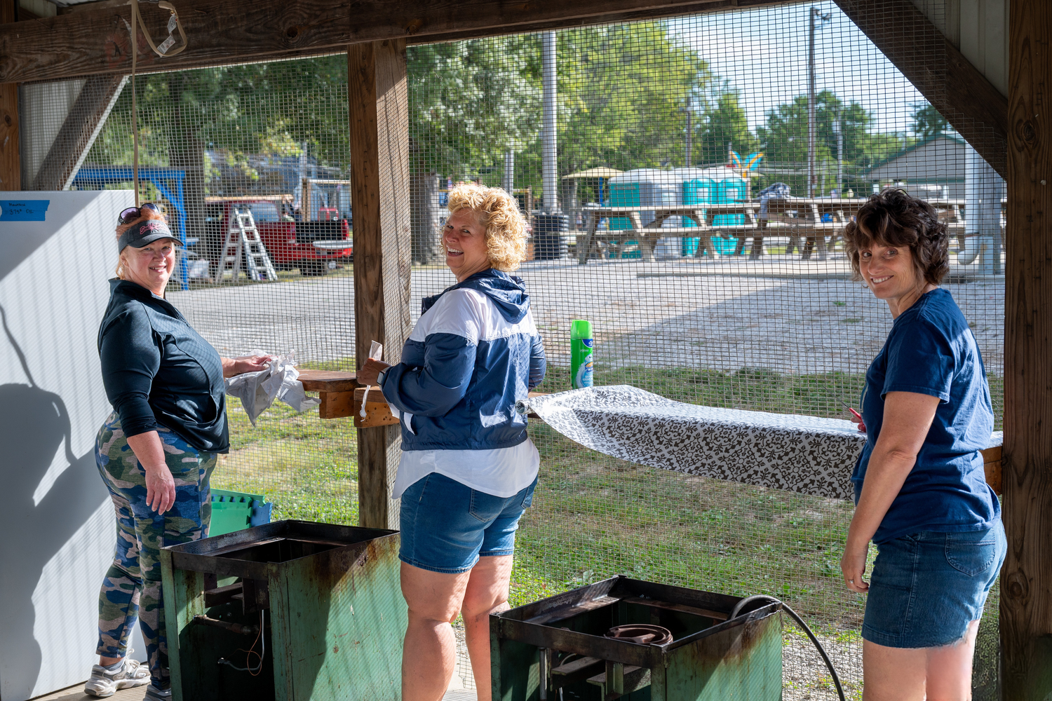 Three women standing under a covered outdoor workspace at a recreational facility on a sunny day.