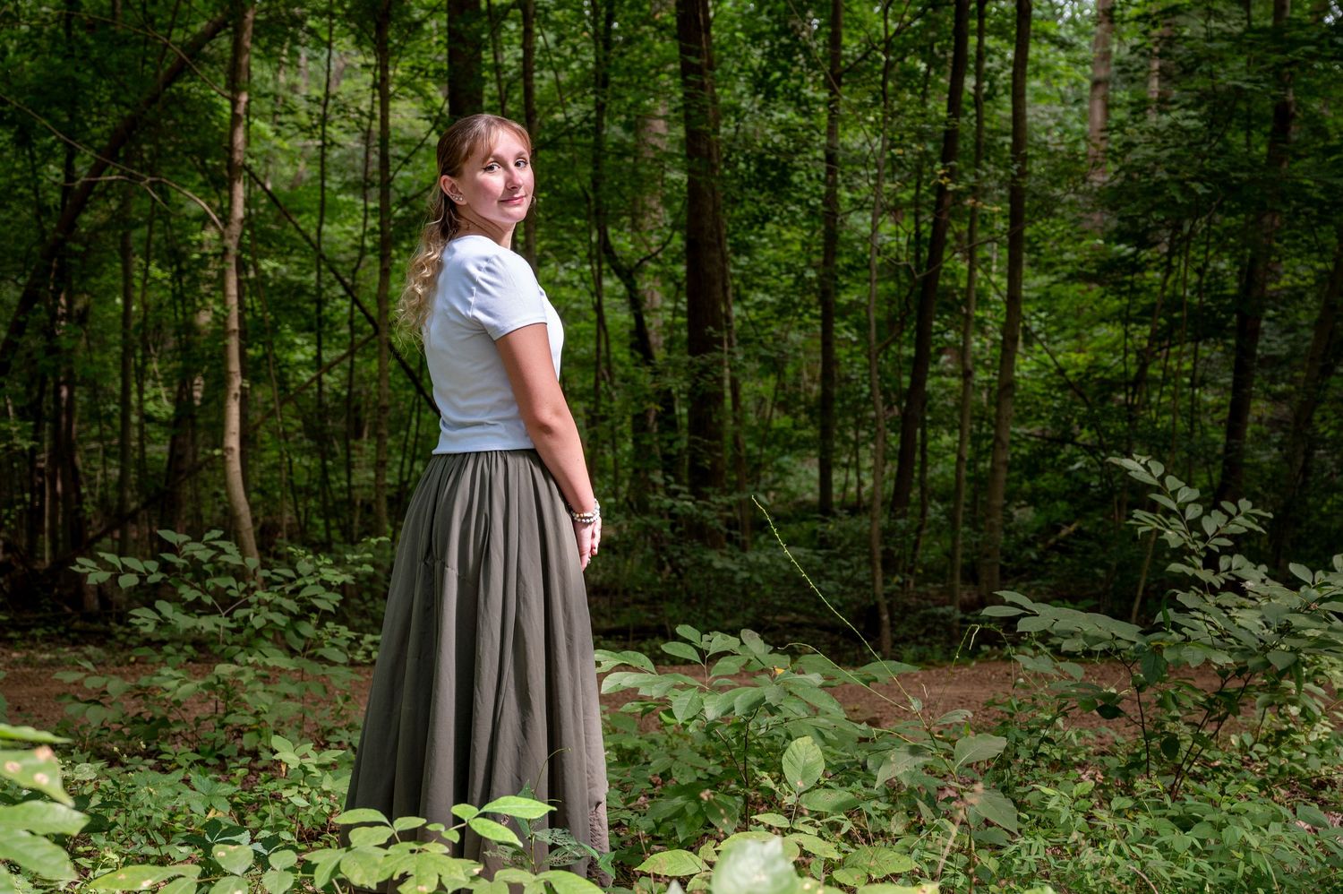 Woman in gray skirt and white top poses in lush green forest.