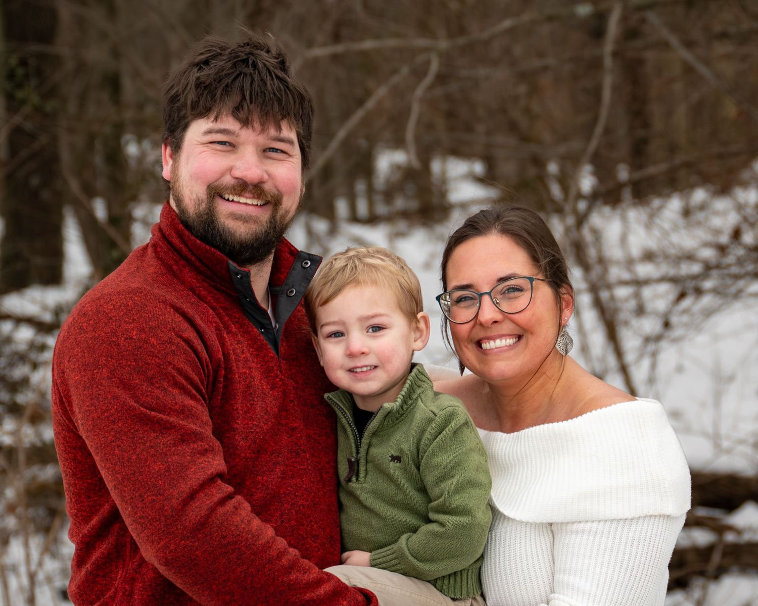 Happy family of three with young toddler boy posing together in snowy winter outdoor setting.