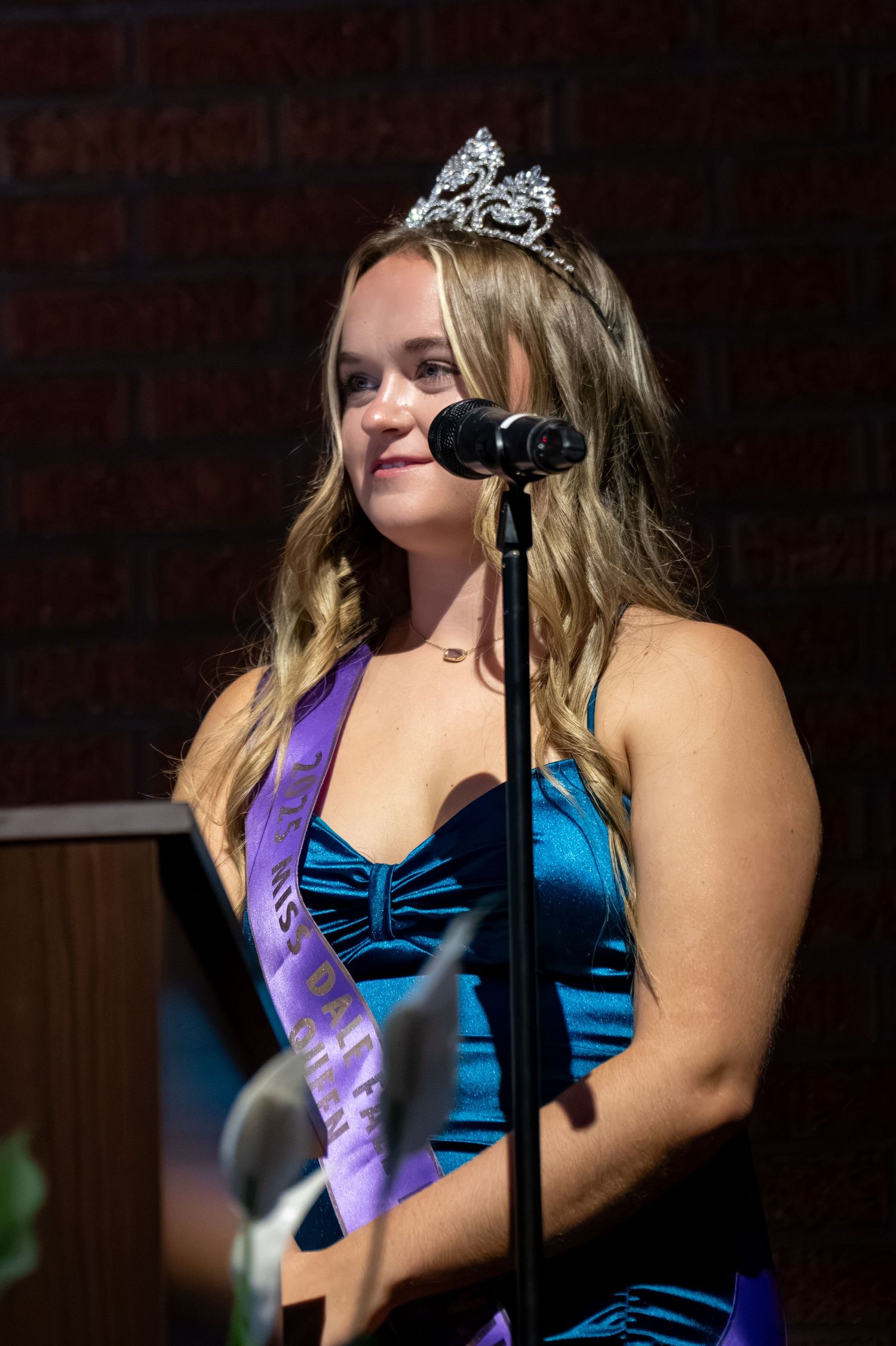 Young woman wearing a tiara and purple sash speaks at a microphone in a blue gown at a pageant event.