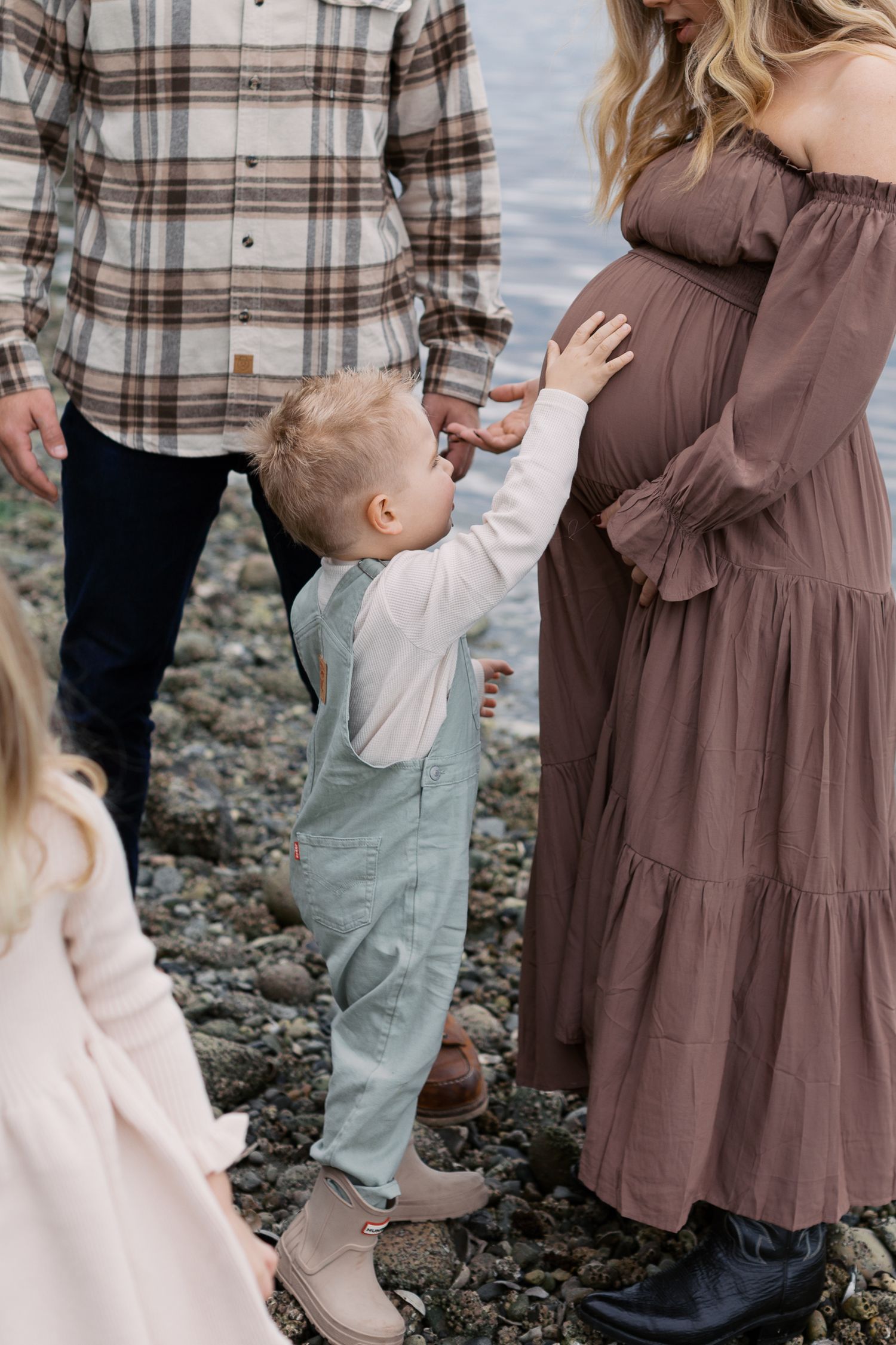 A young boy touches a pregnant woman's belly outdoors near a rocky shore during a family maternity photo session.
