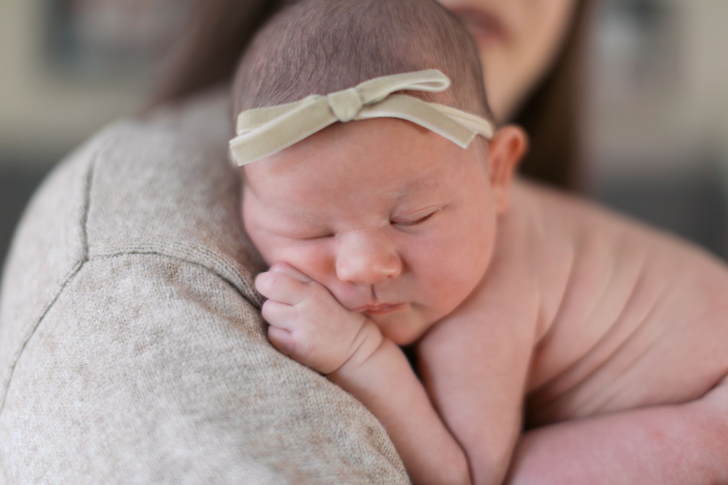 Newborn baby sleeping peacefully on mother's shoulder, wearing a delicate cream bow headband.