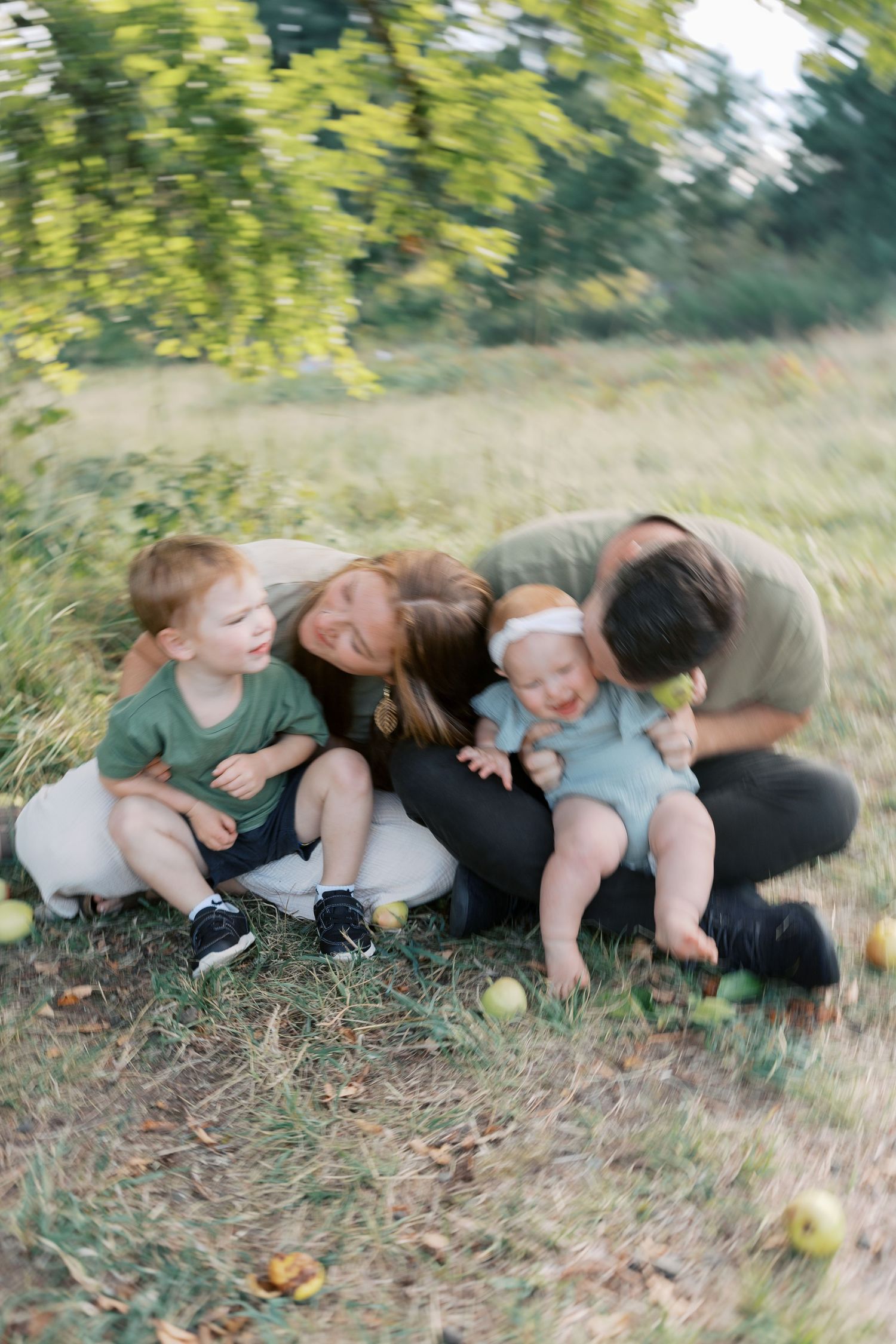 A happy family with two young children sitting together on grass in a lush green park, sharing a joyful moment outdoors.