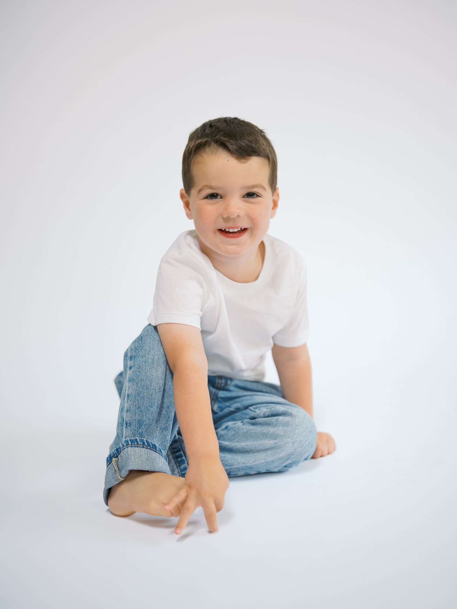 A smiling young boy in a white t-shirt and jeans sits cross-legged on a white background.