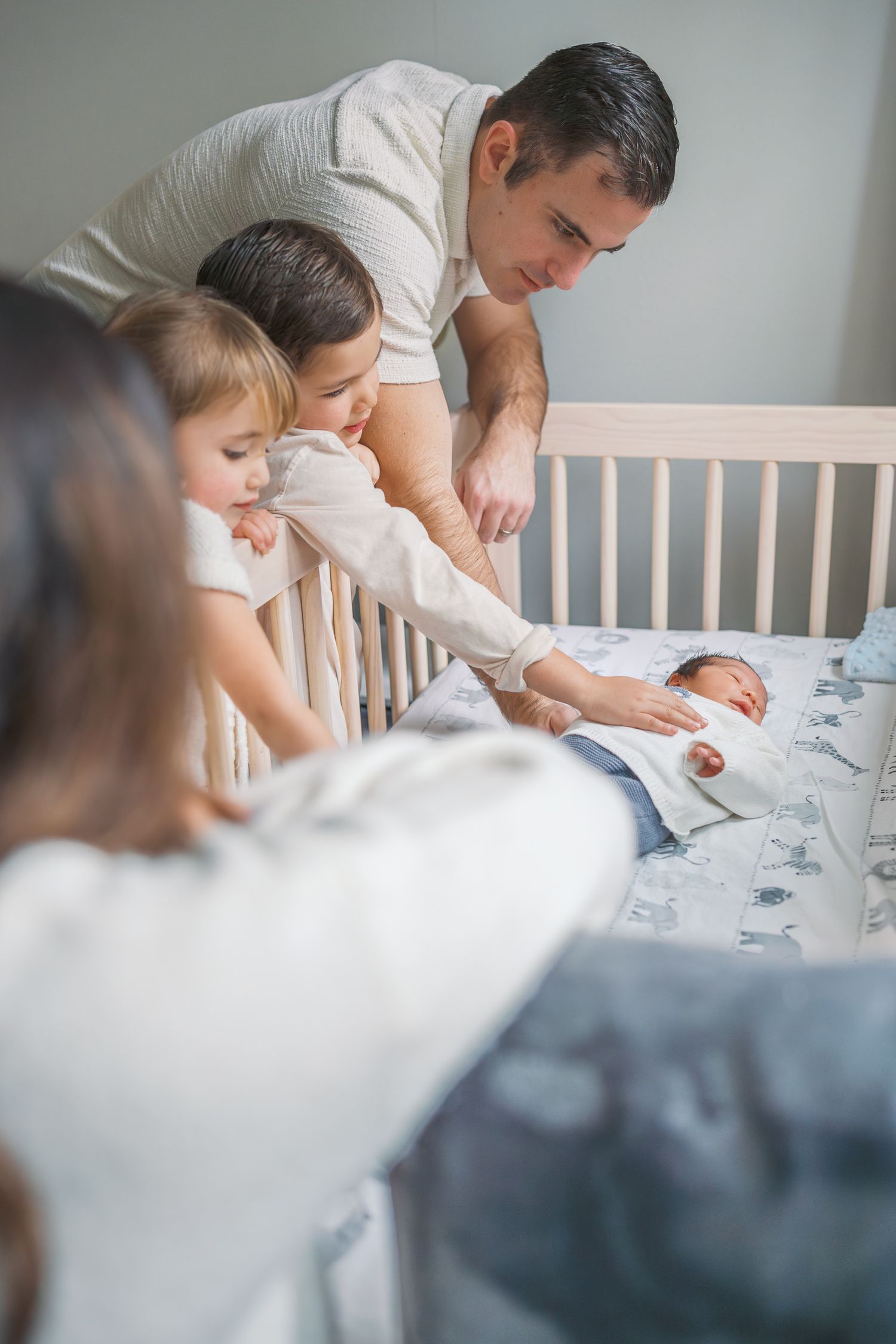 Family with toddler and father leaning over white crib to look at newborn baby lying on patterned sheets.