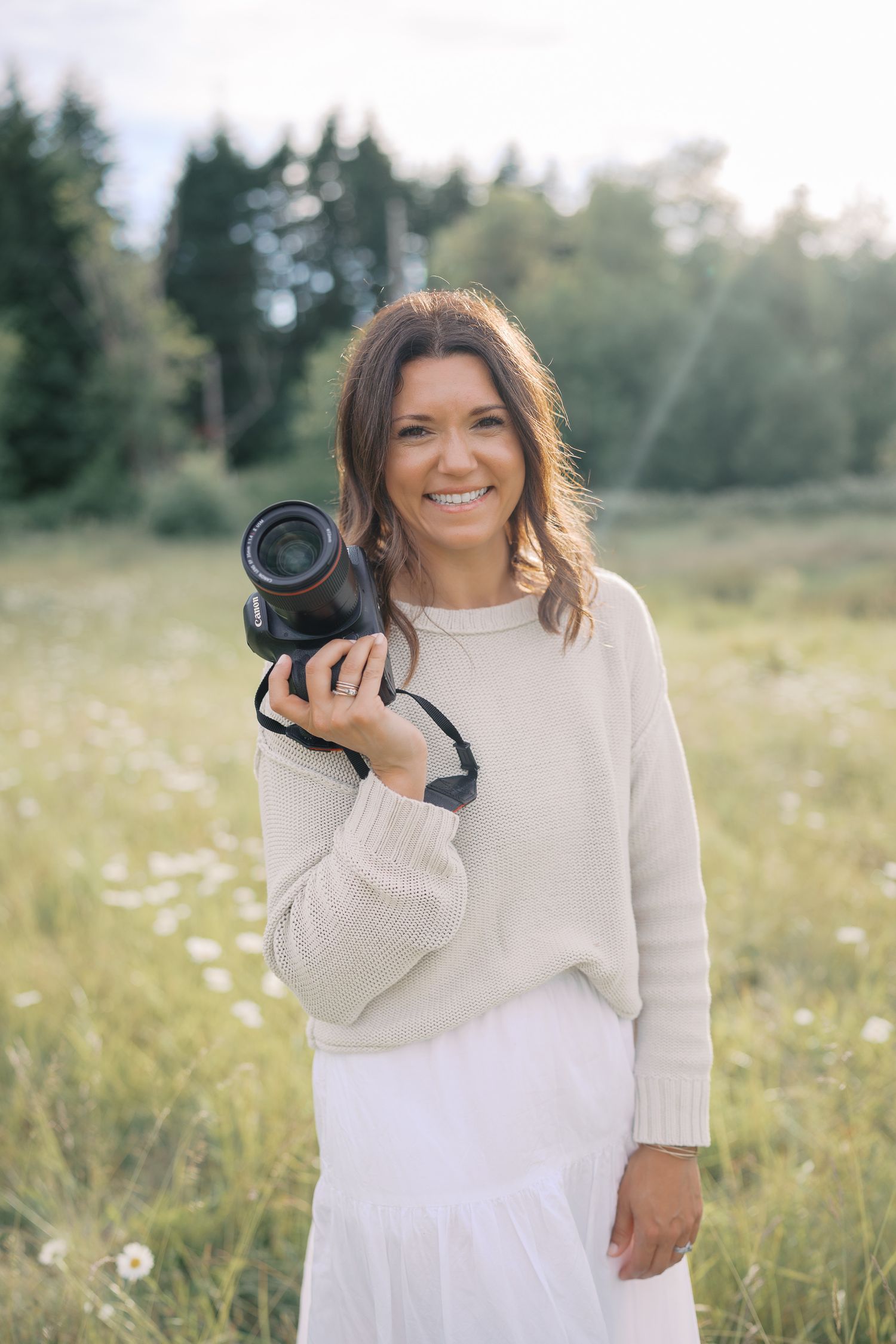 Smiling woman holding a camera in a sunlit meadow, wearing a cream sweater and white skirt.