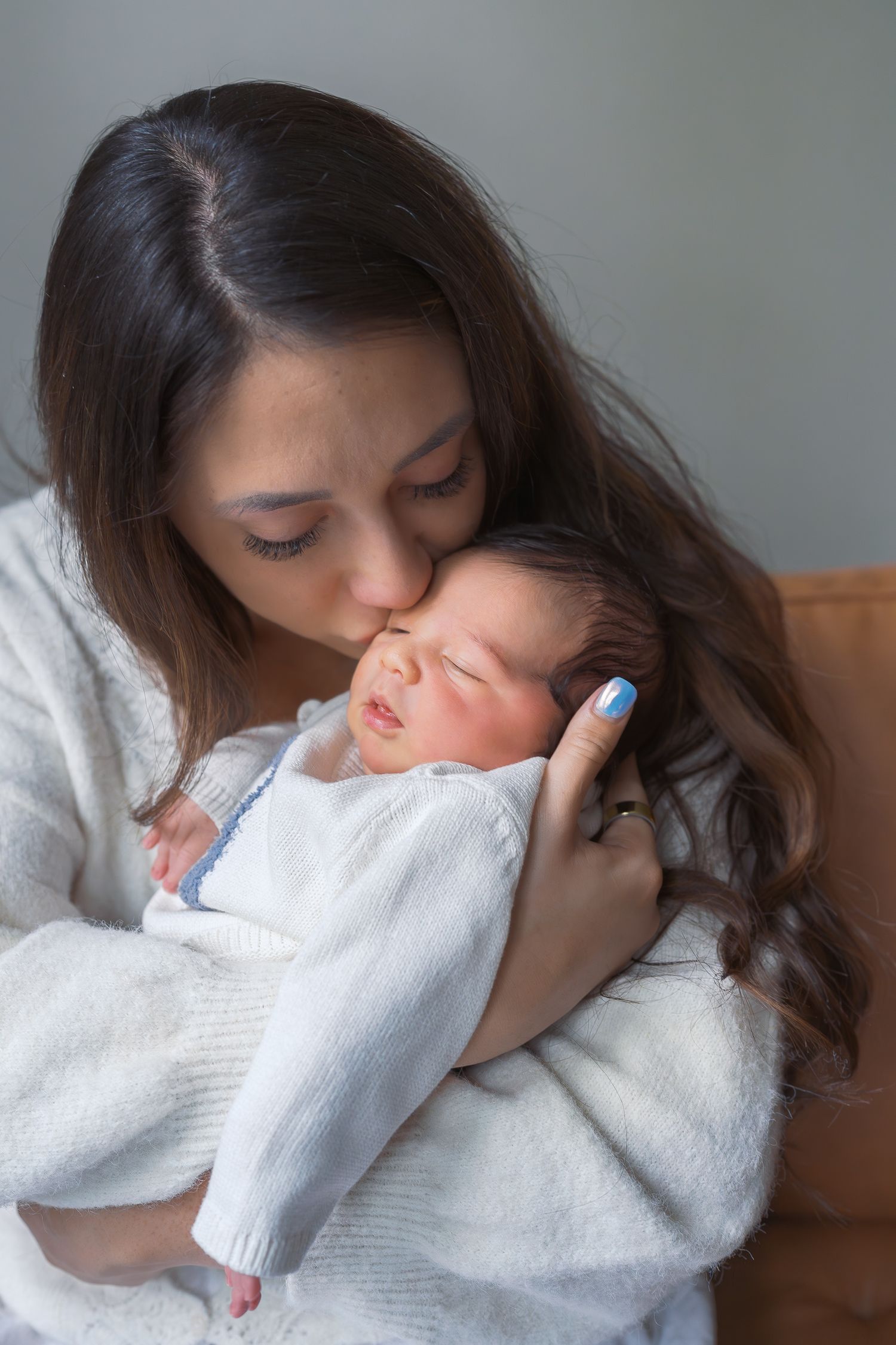 A mother tenderly kisses her sleeping newborn baby wrapped in a white blanket.