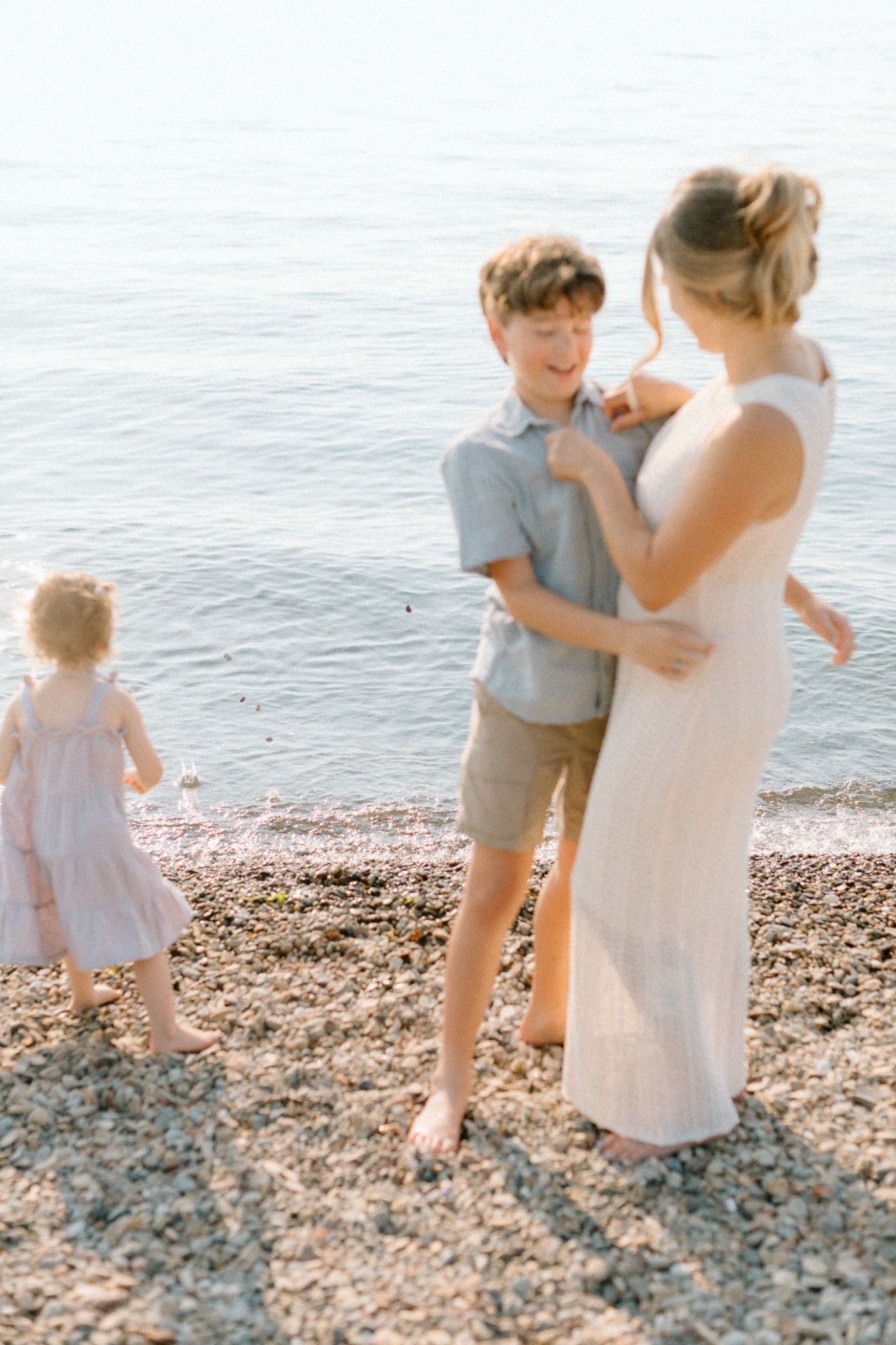 A mother and son share a tender moment on a rocky beach while a young girl in a pink dress plays near the water.