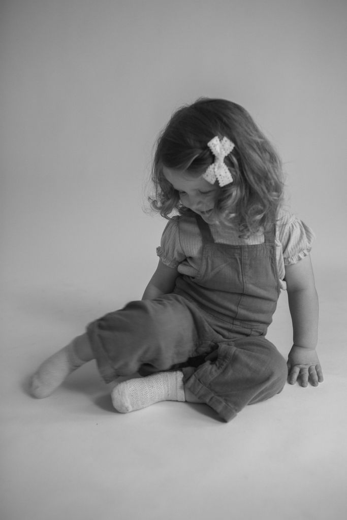 Black and white photo of a young girl in overalls and hair bow, sitting on the floor looking down.