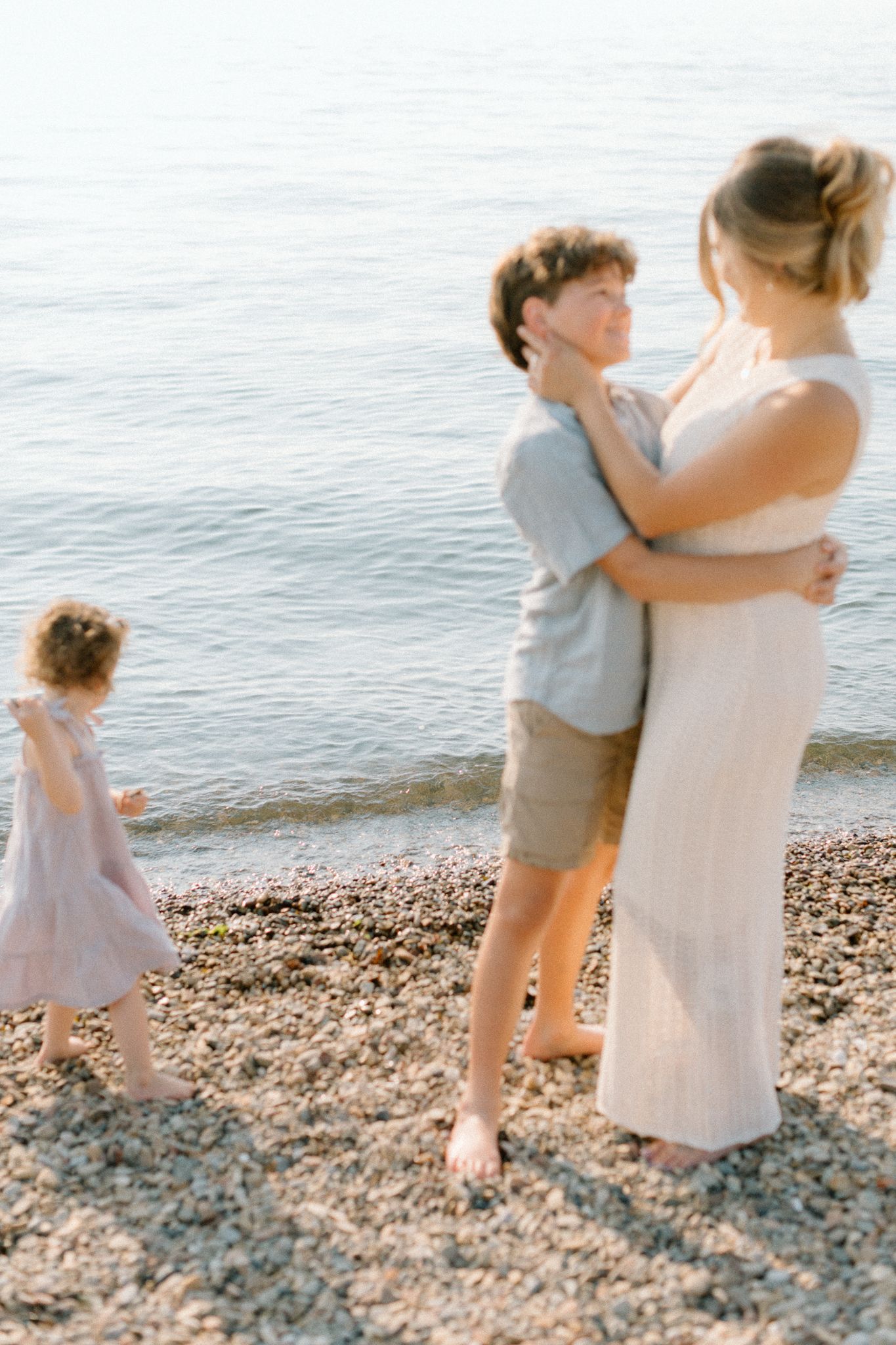Family enjoying a peaceful moment on a pebble beach, with two adults embracing and a young child playing near the water.