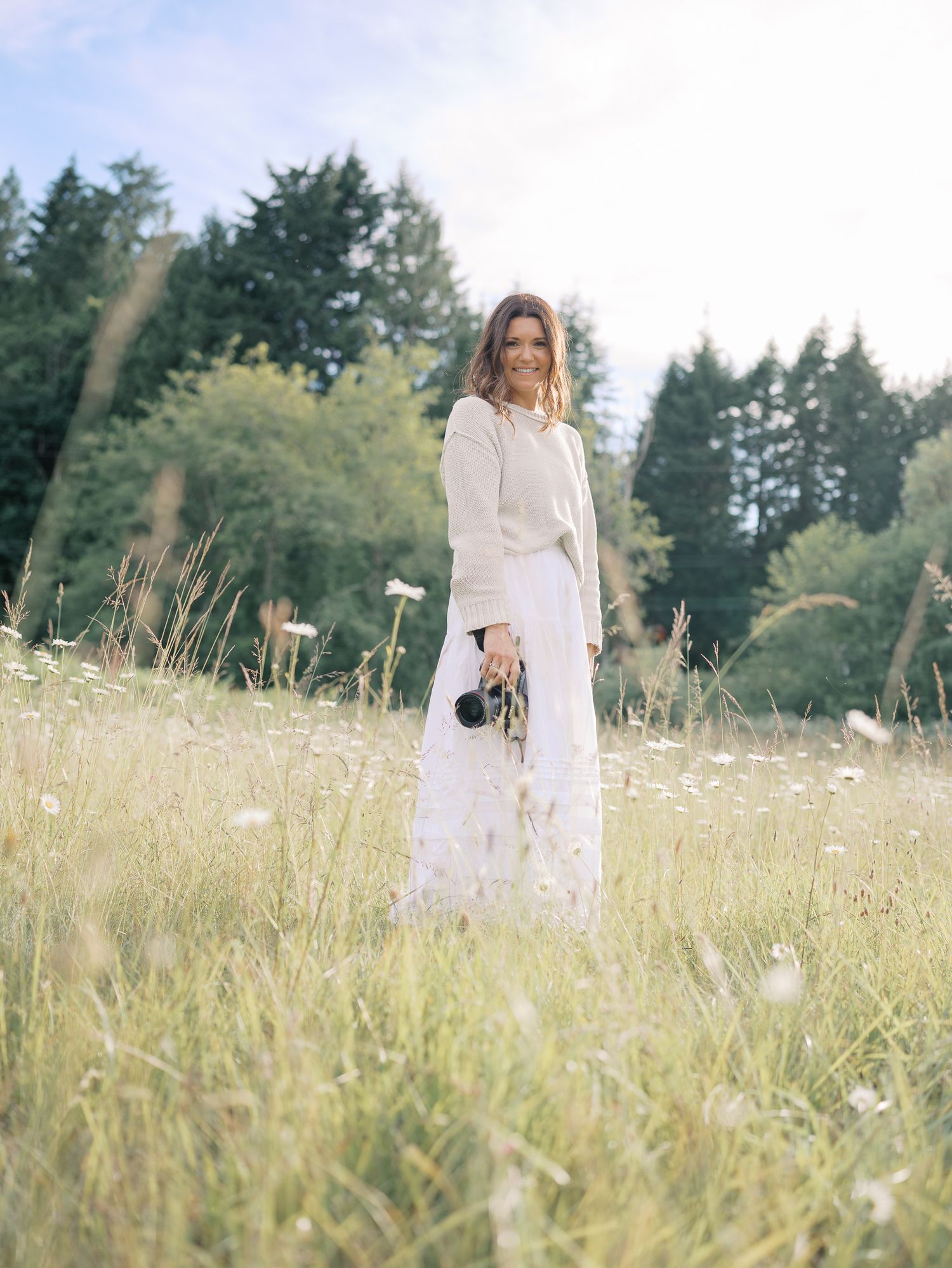 Woman in white outfit holding camera standing in a sunlit meadow with wildflowers and tall trees in background.