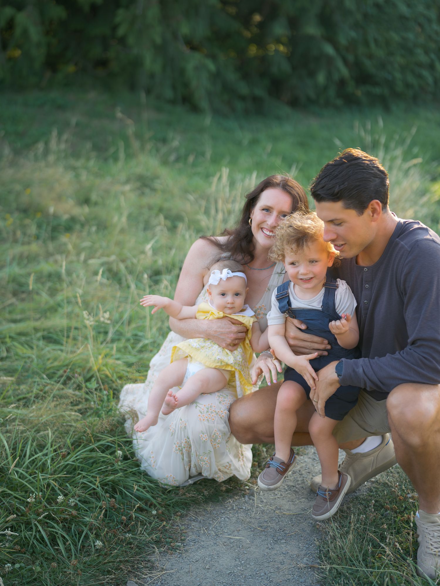 A young family with two toddlers smiling together outdoors in a lush green meadow during golden hour.