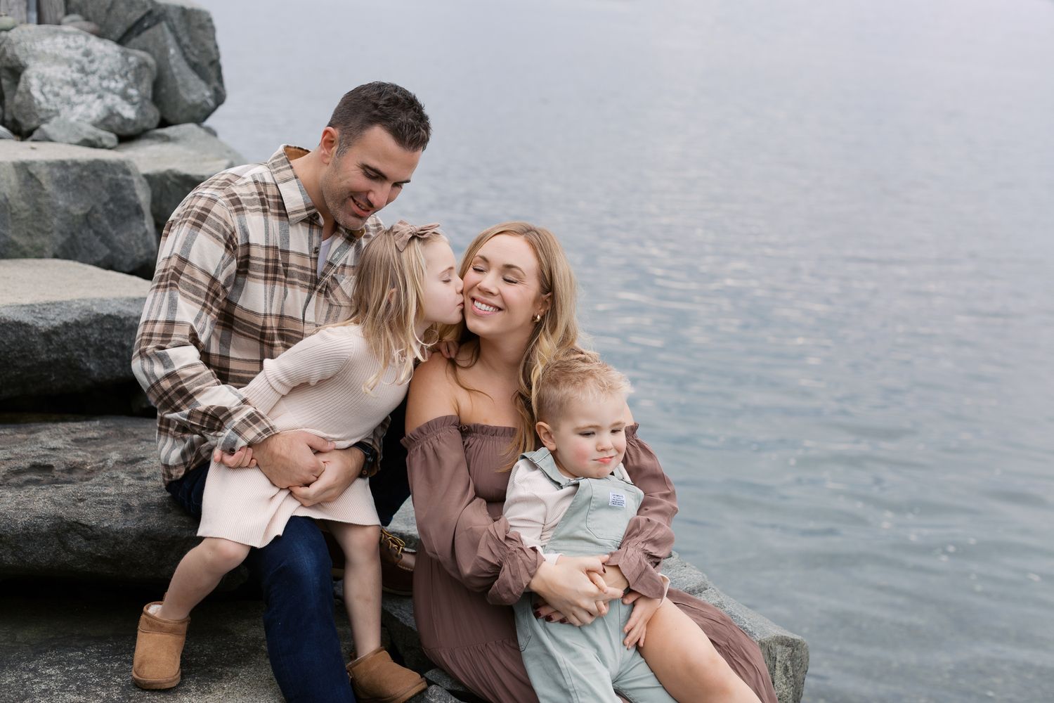 Happy family of four sitting on rocky waterfront, parents smiling with young children in casual outdoor portrait.
