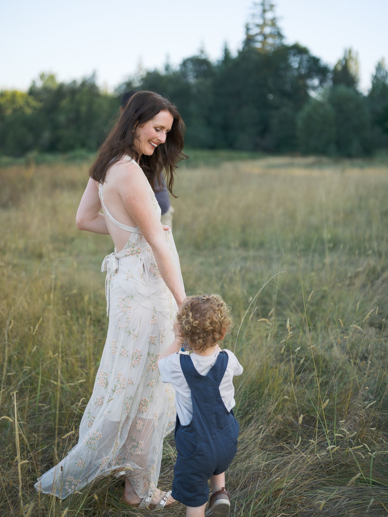 A mother in a lace dress holds hands with her toddler in overalls in a sunlit meadow.