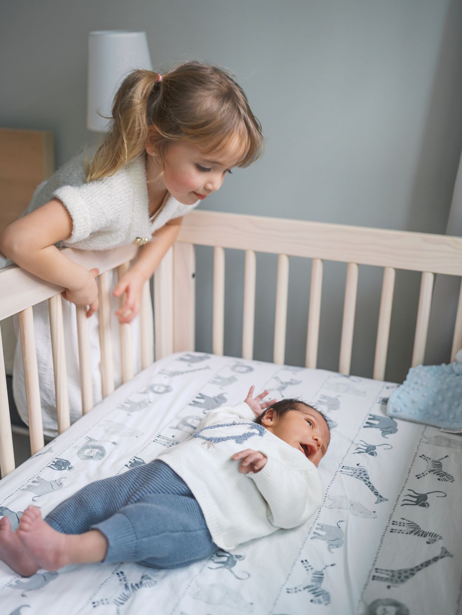 A toddler girl leans over a wooden crib, gazing lovingly at a newborn baby lying on a animal-print blanket.