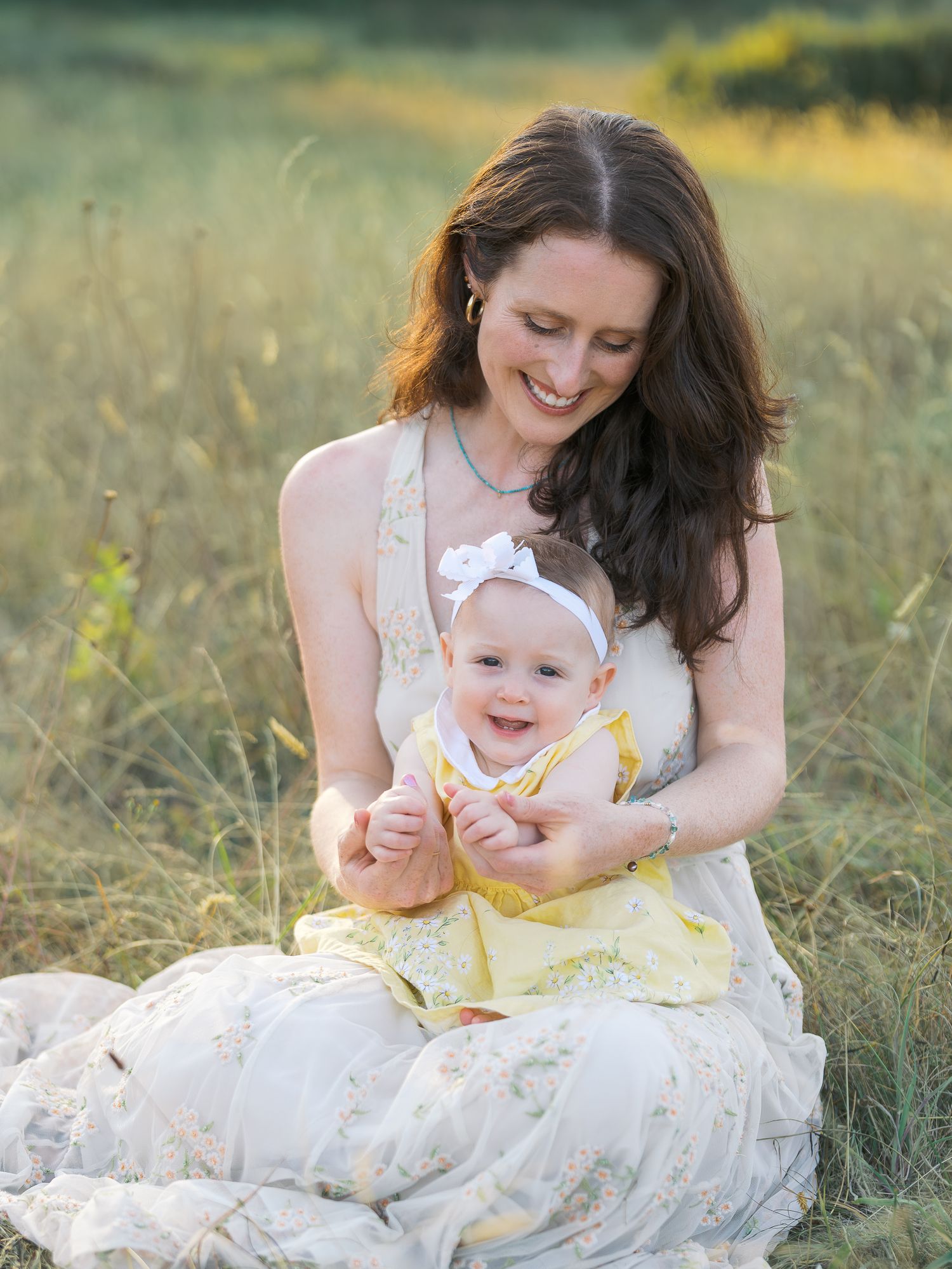 A smiling mother sits in a sunny meadow holding her happy baby girl in a yellow dress with a white bow headband.