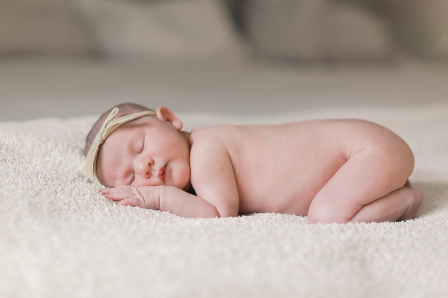 Peaceful newborn baby sleeping curled up on a soft white blanket wearing a delicate headband.