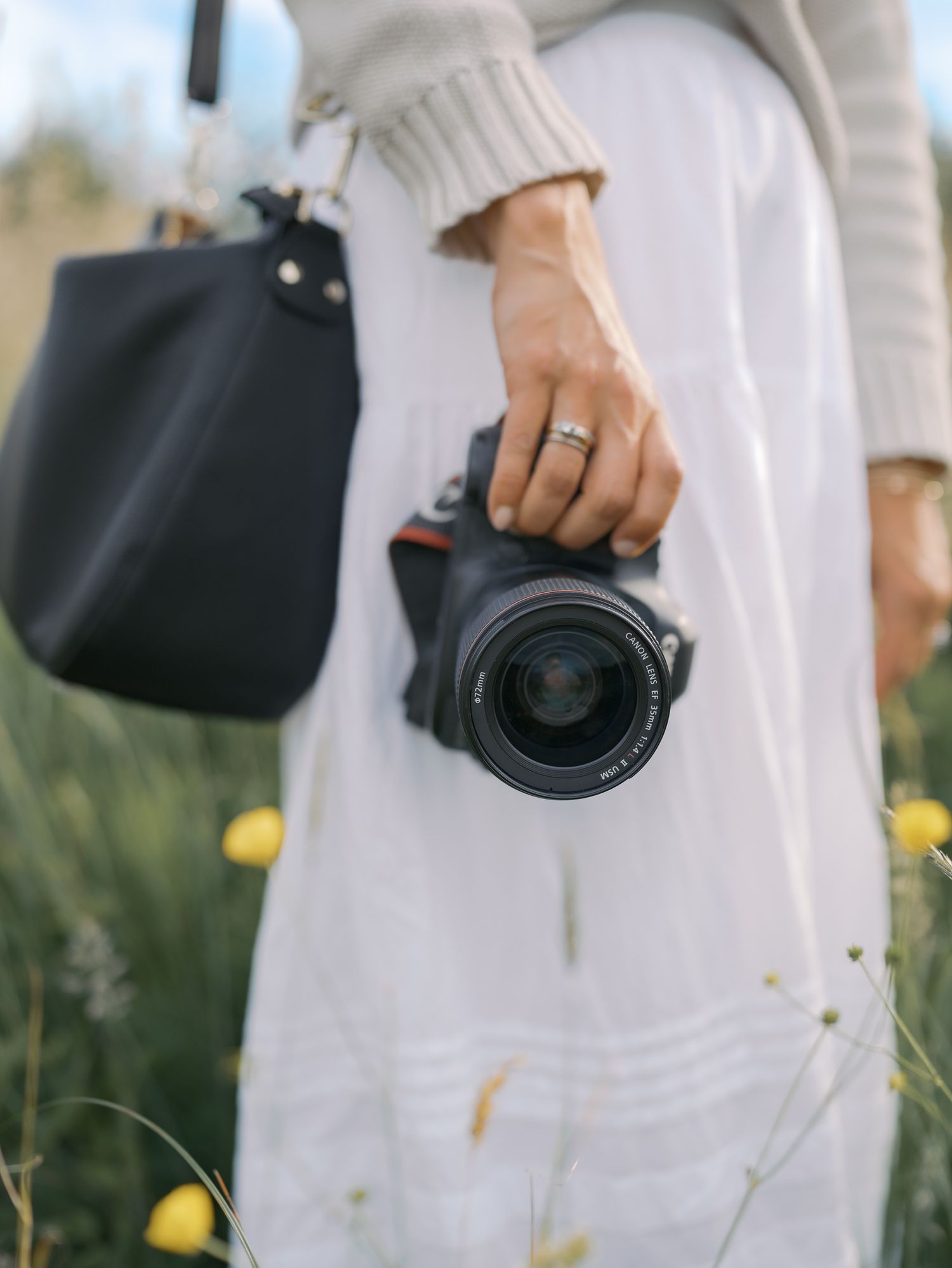 Woman in white dress holding a DSLR camera and black leather bag while standing in a wildflower field.