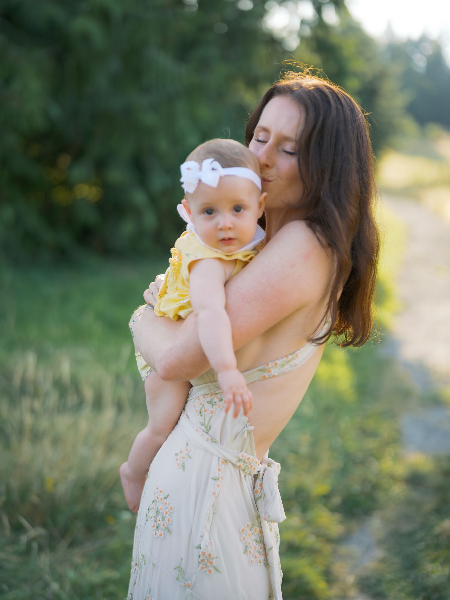 A mother tenderly kisses her baby girl in a yellow dress outdoors in golden hour light.