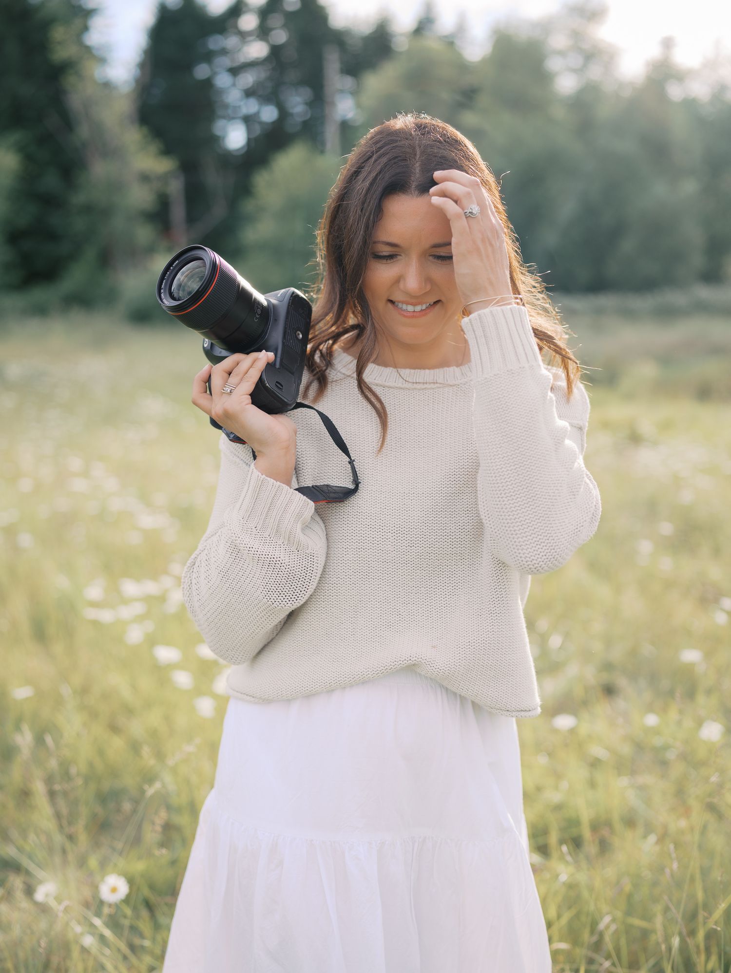 Woman in beige sweater and white skirt holding a DSLR camera while standing in a sunny wildflower field.