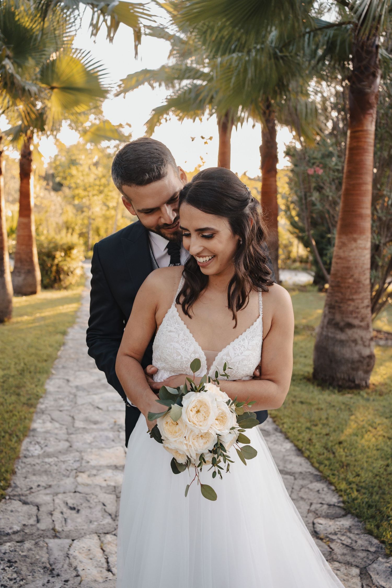 Newlyweds embrace under palm trees with bride holding white rose bouquet.