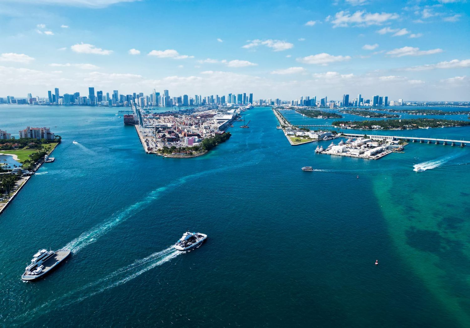 Aerial panoramic view of Miami skyline with turquoise ocean waters, boats, and downtown buildings under blue skies.
