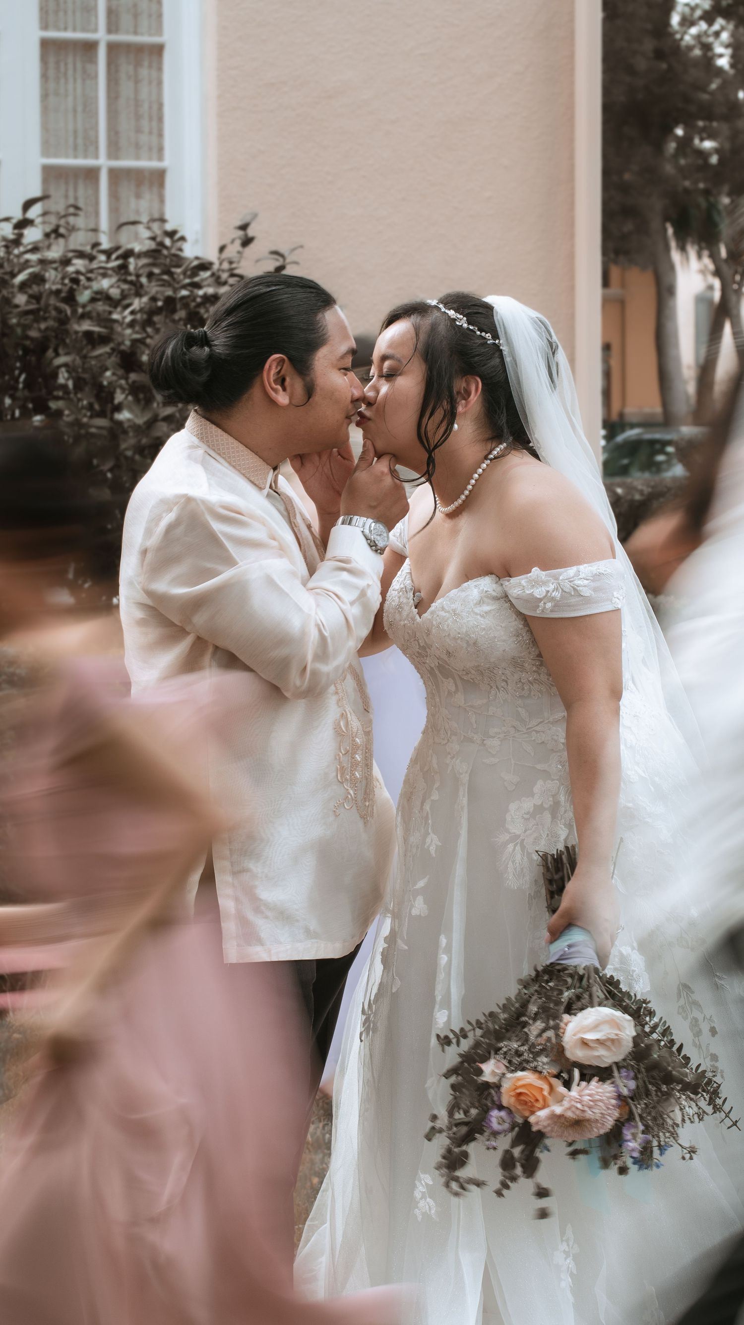 Romantic wedding moment captured as newlywed couple shares tender kiss outdoors with floral bouquet in foreground.